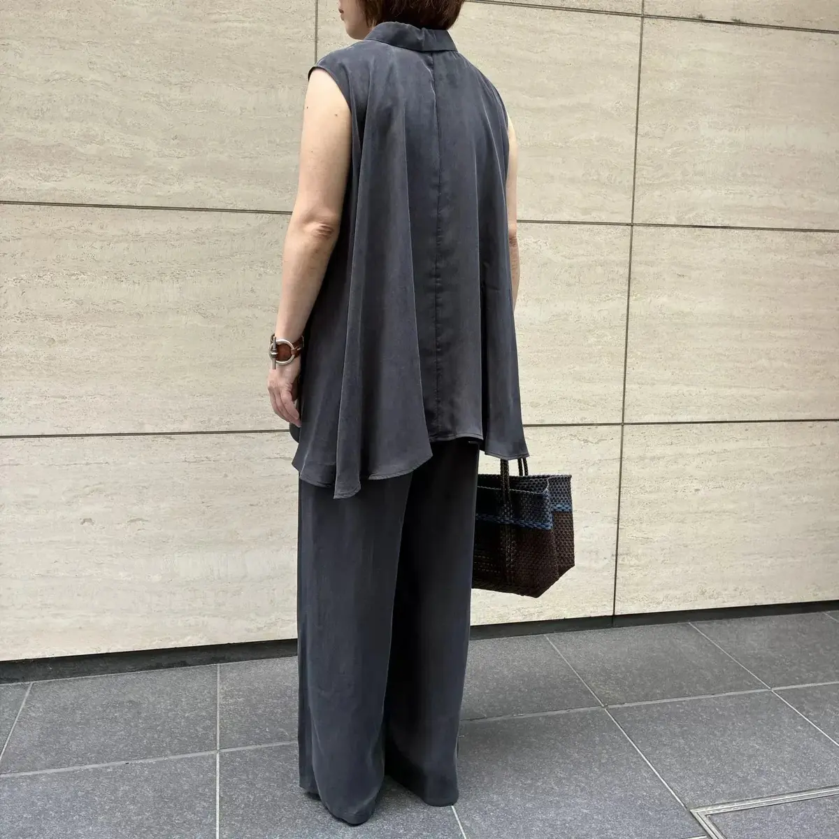 グレーのスリーピース。襟付きのノースリーブトップスとワイドパンツ、そしてロングカーディガン。女性が後ろを向いて立っており、手には編み込み模様のバッグを持っている。背景はベージュのタイル壁。