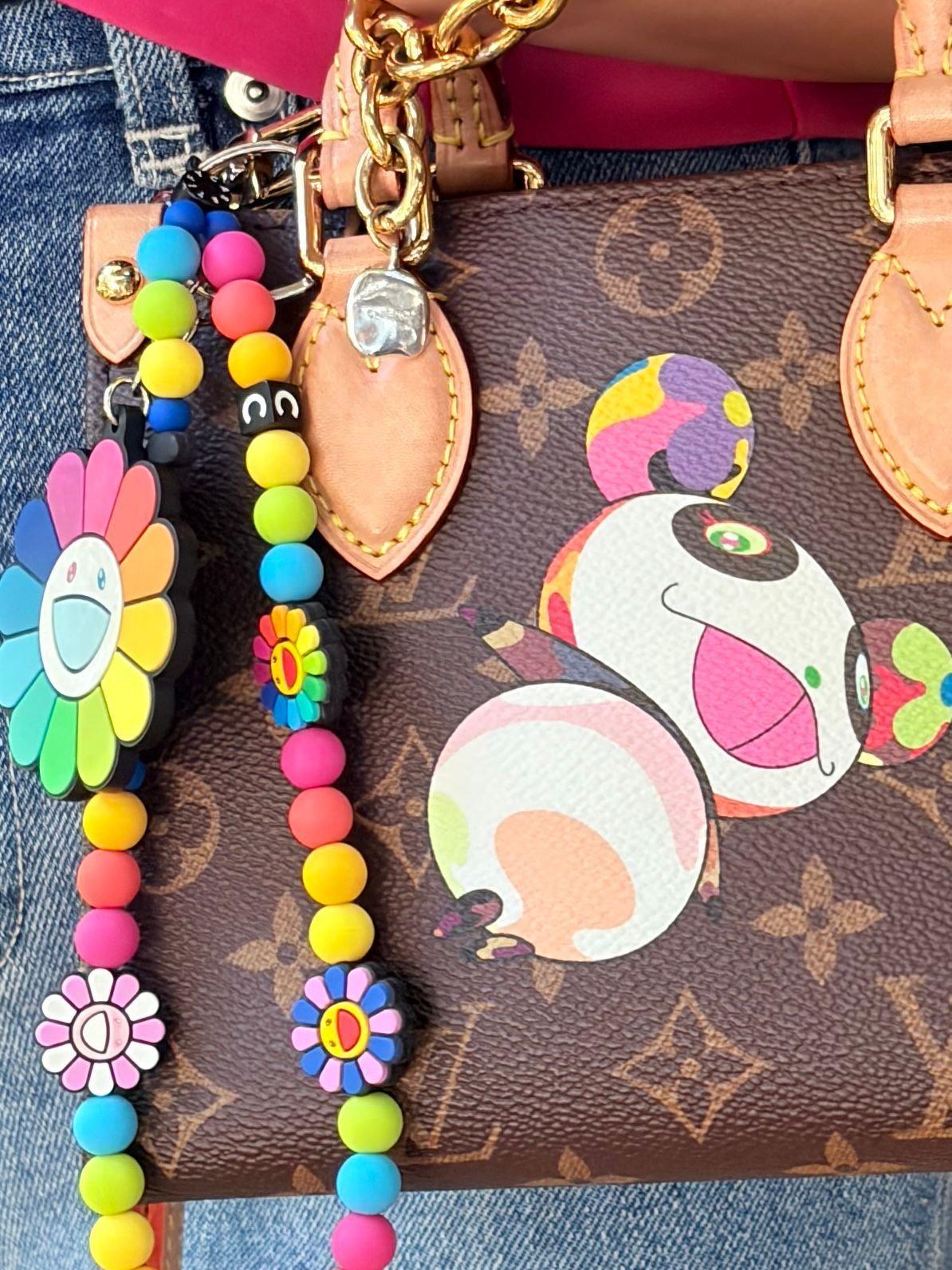 ルイ・ヴィトンのバッグに、カラフルな花のモチーフのキーホルダーが取り付けられている。バッグには、白とピンクの目をしたキャラクターのイラストが描かれている。