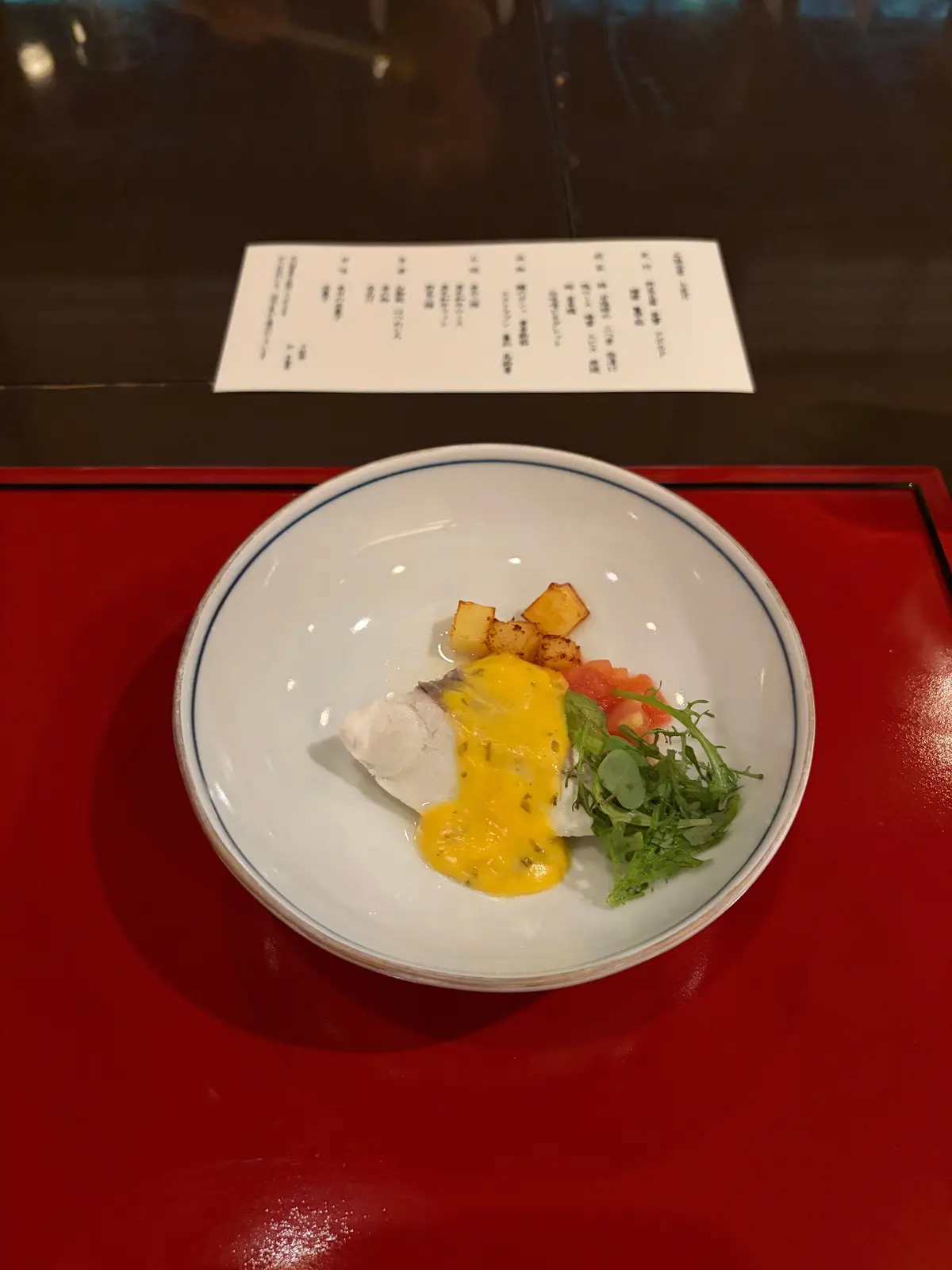 青い皿に盛り付けられた料理。白身魚、ポテトの角切り、黄色いソース、トマトとハーブの彩りが特徴。背景にはメニューが置かれている。