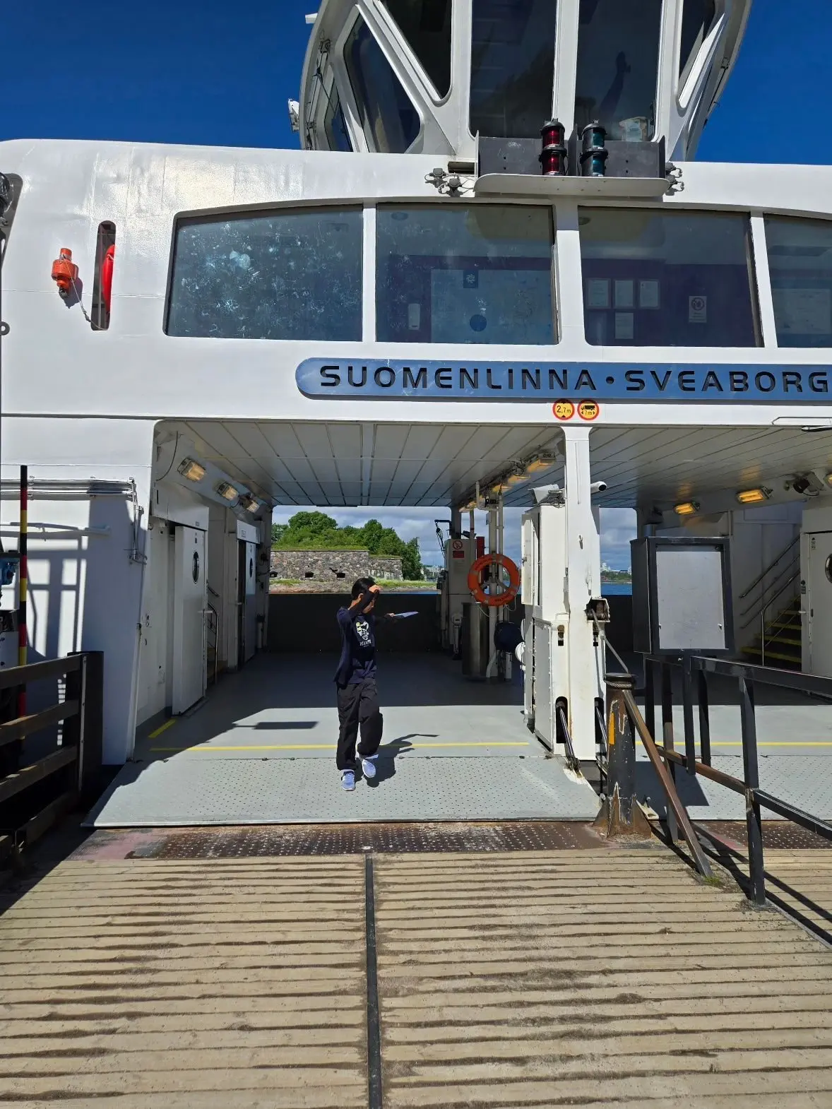 白船の船首部分。青い文字で「SUOMENLINNA・SVEABORG」と表示。中央に人が立っており、背景に水辺の風景が広がる。