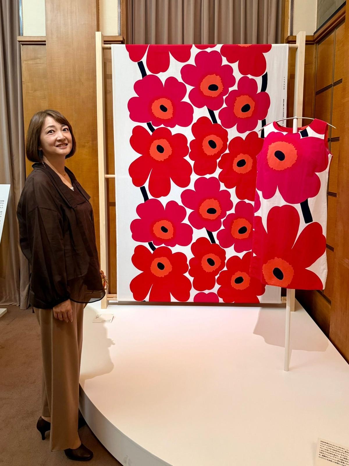 展示ケースに飾られた、赤とピンクの大きなポピーの花柄の白い布。女性が隣に立って微笑んでいる。背景には木製の壁とカーテンが見える。
