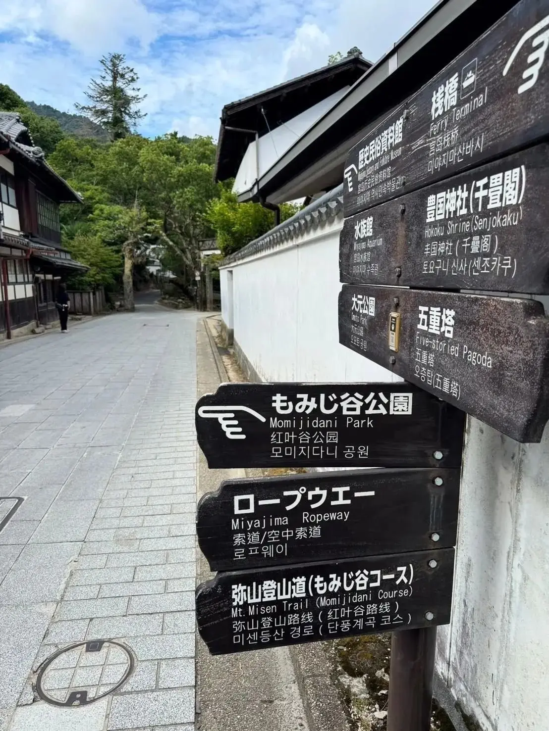 宮島の案内板。木製の看板に、日本語と韓国語で観光スポット（厳島神社、ロープウェイ、弥山登山コースなど）が記載されている。背景には、苔むした石畳の道と伝統的な建物が見える。