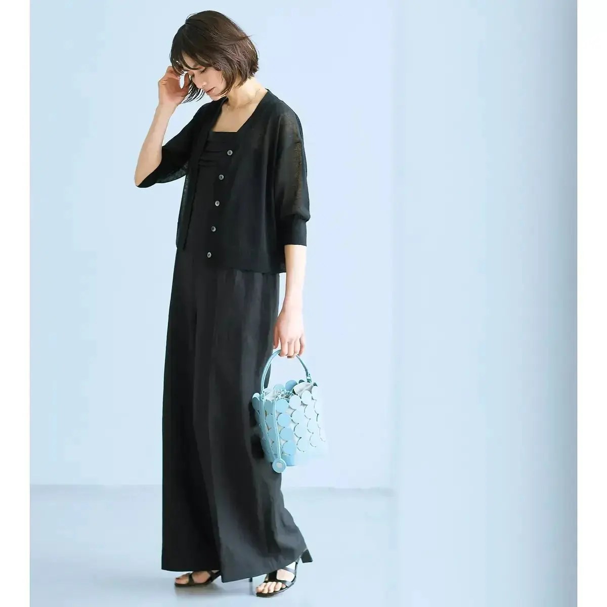 女性が黒の長袖チュニックとロングスカートを着用し、軽い手袋を片手に持つ。スカートにはボタン装飾があり、足元は黒のサンダル。右手には青い円形模様のバスケットを所持。背景は無地で、全体がシンプルなデザイン。