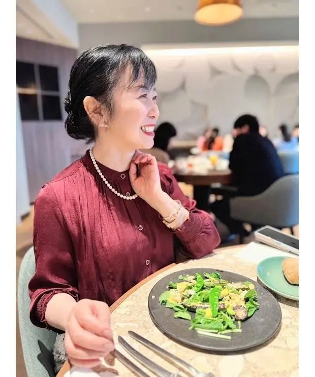 レストランで食事を楽しむ女性。赤いブラウスを着用し、パールネックレスを身につけている。笑顔でこちらを見ている。テーブルにはサラダとパンが置かれている。背景には他の客や装飾が見える。