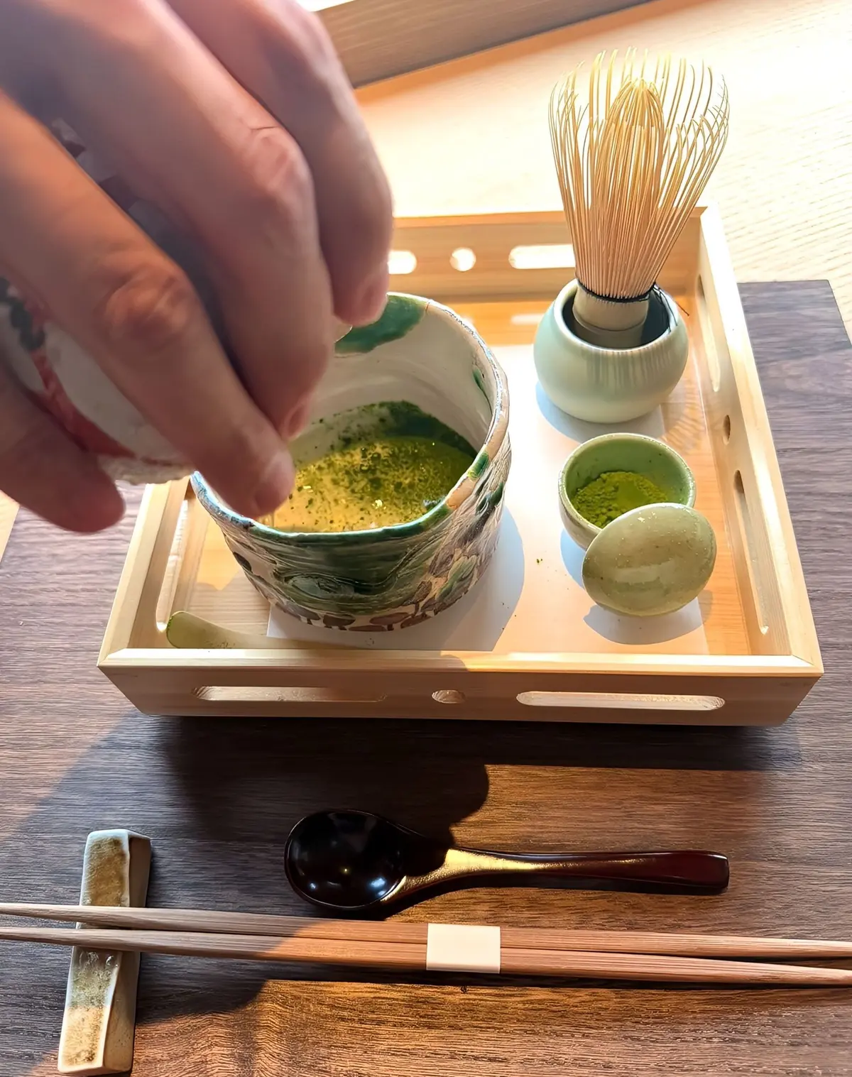 木製のトレイに抹茶、茶碗、茶筅、小分け容器が置かれている。手で抹茶を茶碗に振り入れている様子。茶碗は白地に緑の模様。