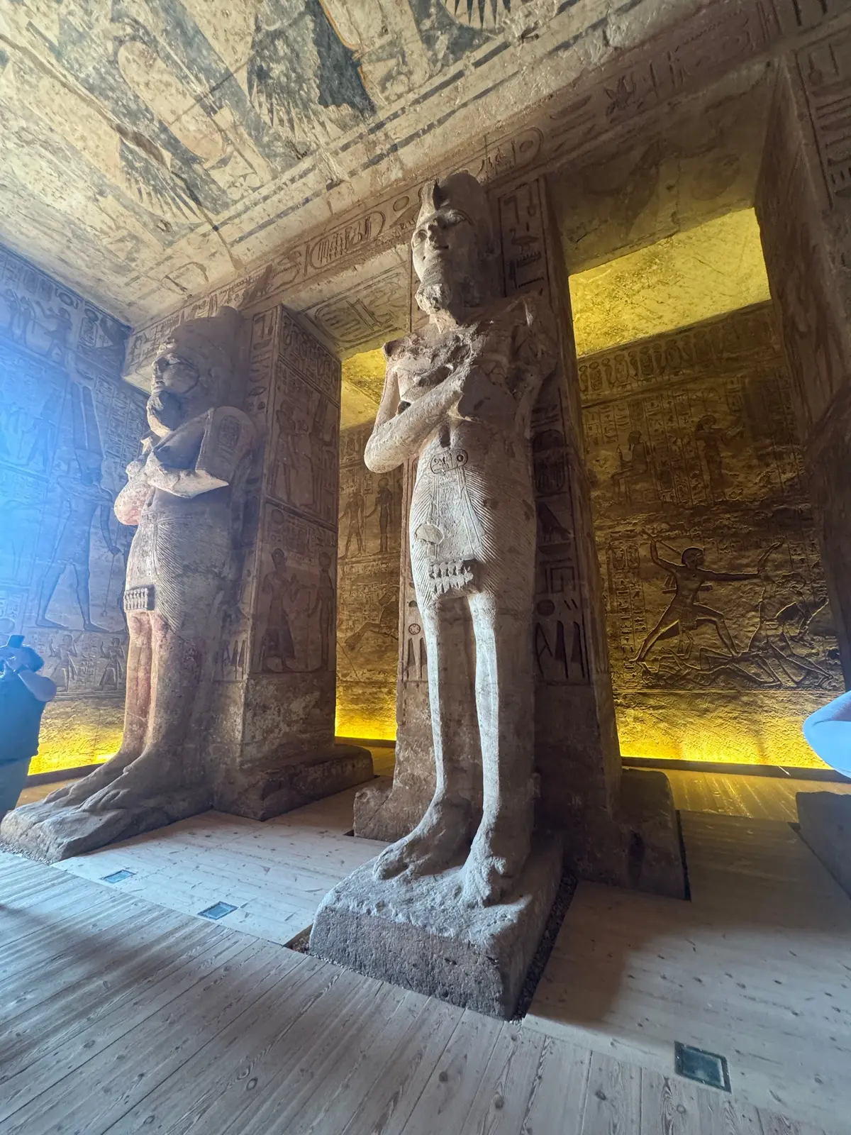 古代エジプトの壁画が彫刻された岩壁の内部。中央には神々らしき人物が描かれ、上部にはヒエログリフが並ぶ。女性が壁画を眺めている。
