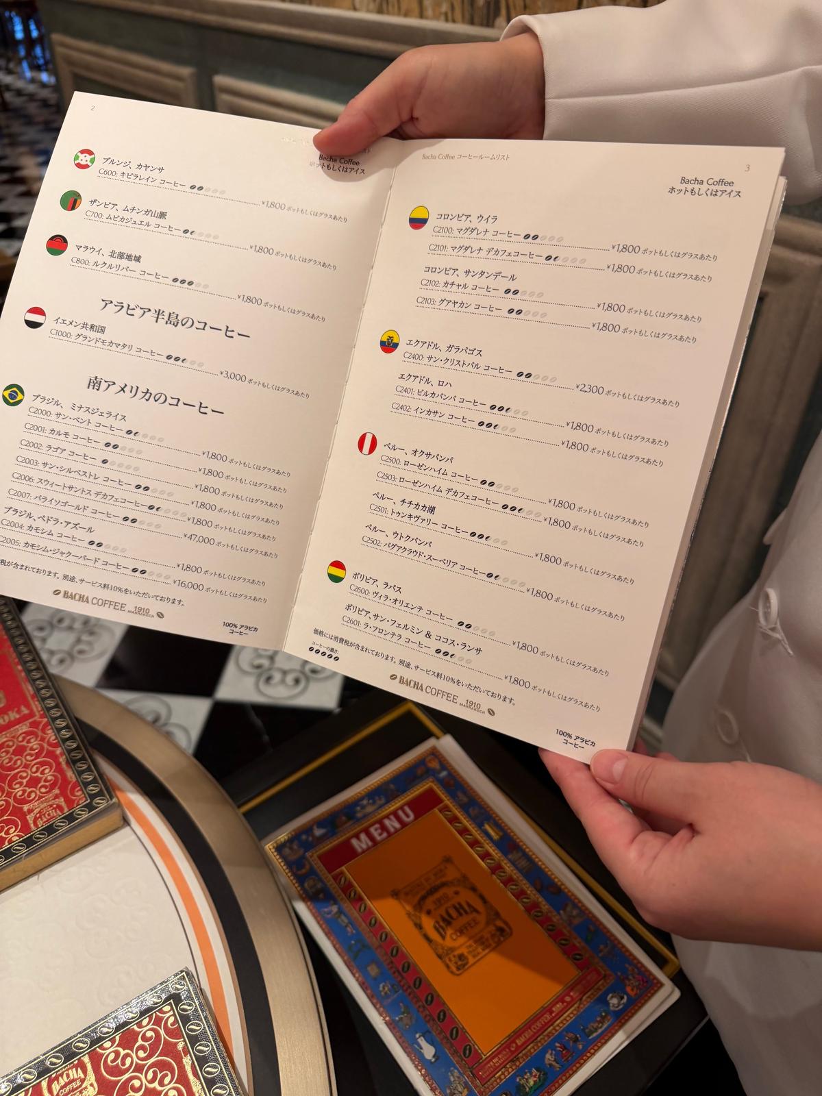 カフェのメニューが記載されたA4サイズの紙。左上には「Racha Coffee」のロゴマークがあり、右ページには「Racha Coffee ホットもアイスも」の文字。メニューにはコーヒーやドリンクの品名、価格、説明が日本語で記載されている。