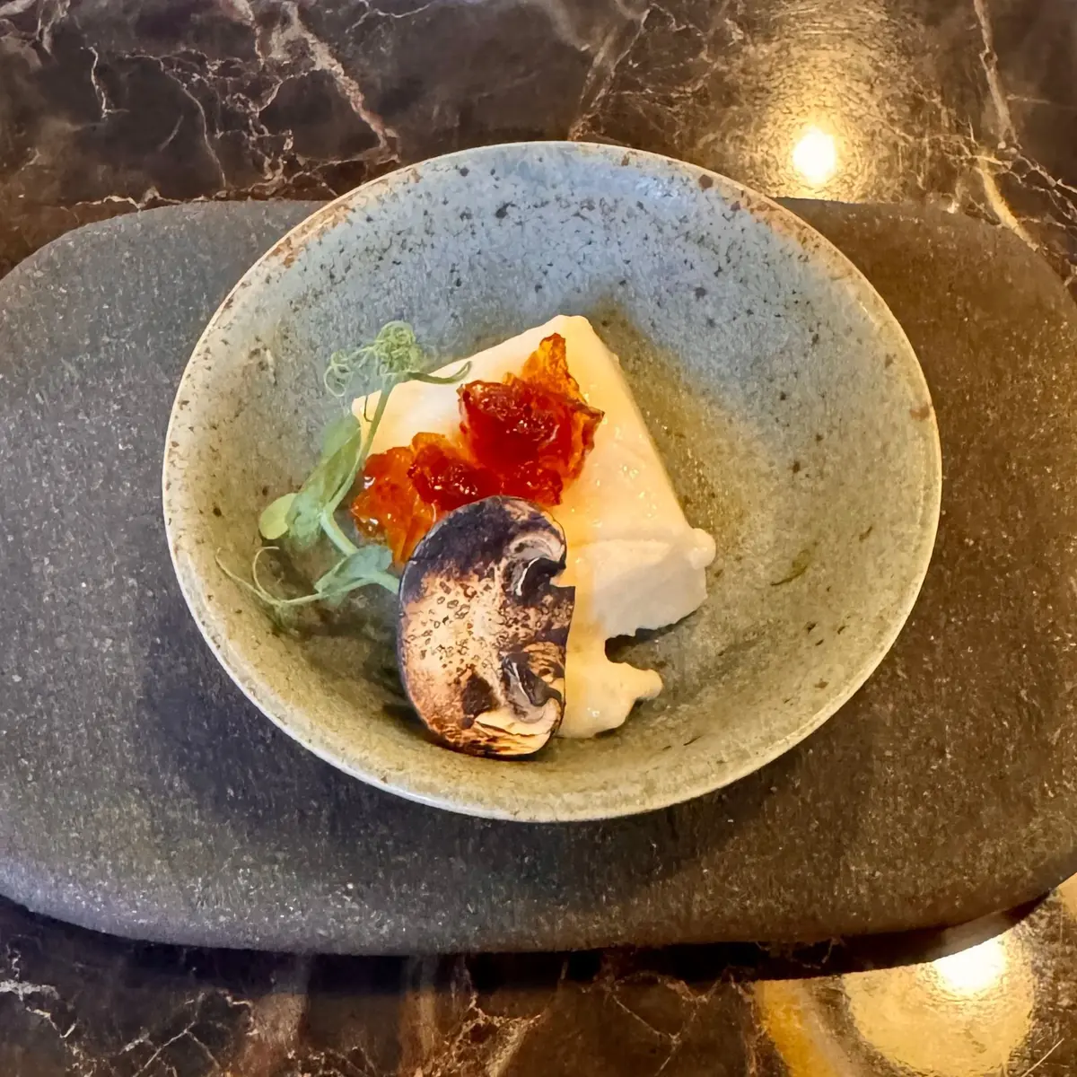 灰色の器に盛り付けられた、白身魚のポワレ。上にオレンジ色の魚卵と、緑の葉、茶色のキノコが添えられている。