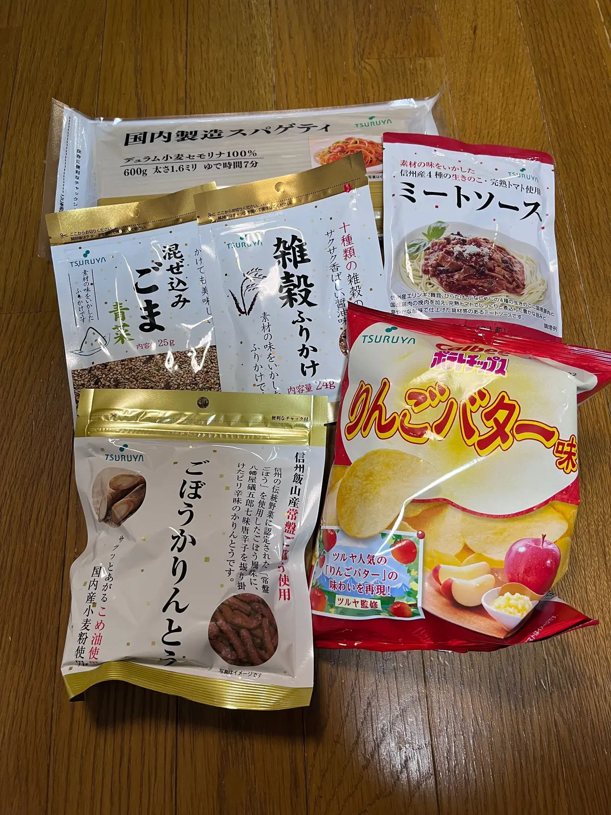 白背景に、様々なツルヤ食品の商品が並んでいる写真。国内製造ラーメン、ミートソース、冷や汁、こんにゃく麺、柿の種など、複数の食品パッケージが横一列に配置されている。カラフルなパッケージが目立ち、右下に「ツルヤ」のロゴが見える。