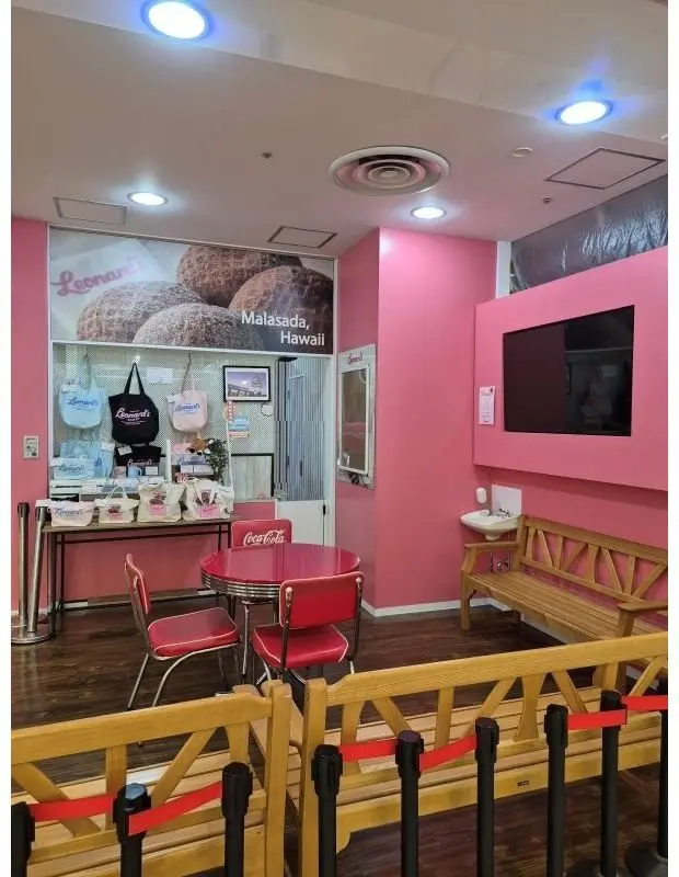 ピンク色の壁に、レナードス（Leonard's）の店舗内。ショーケースにはマラサダが並び、テーブルと赤い椅子が配置されている。右には木製のベンチと洗面台がある。