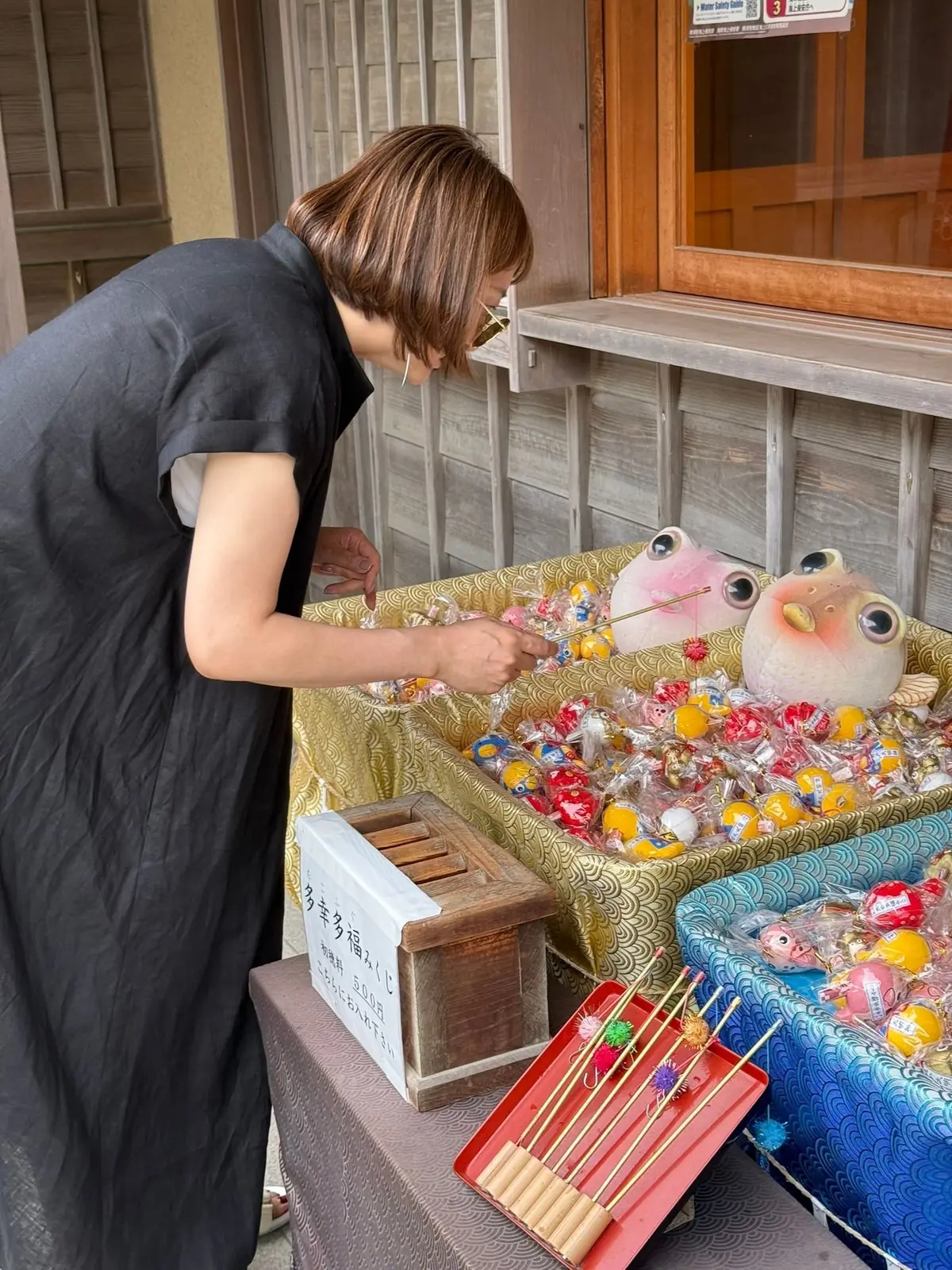黒いワンピースを着た女性が、木製のトレーに置かれたたくさんの人形やぬいぐるみの入った水色の箱に手を伸ばしている。箱の横には募金箱と竹串が置かれている。背景には木造の建物がある。