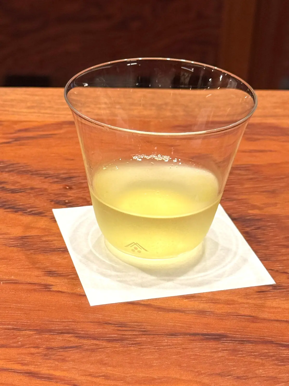 透明なプラスチックカップに淡い黄色の液体が入っており、木製のテーブル上に白いコースターに置かれています。