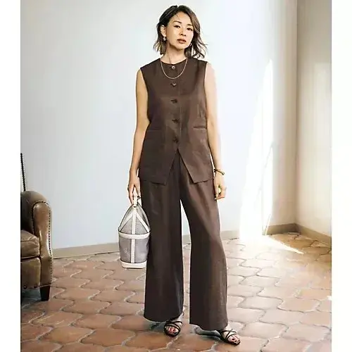 女性が白い壁に寄りかかり、笑顔でポーズしている。白のTシャツに紺のベストを着て、紺のワイドパンツを履き、右手に銀色のクラッチバッグを持っている。足元には白いスニーカーが見える。