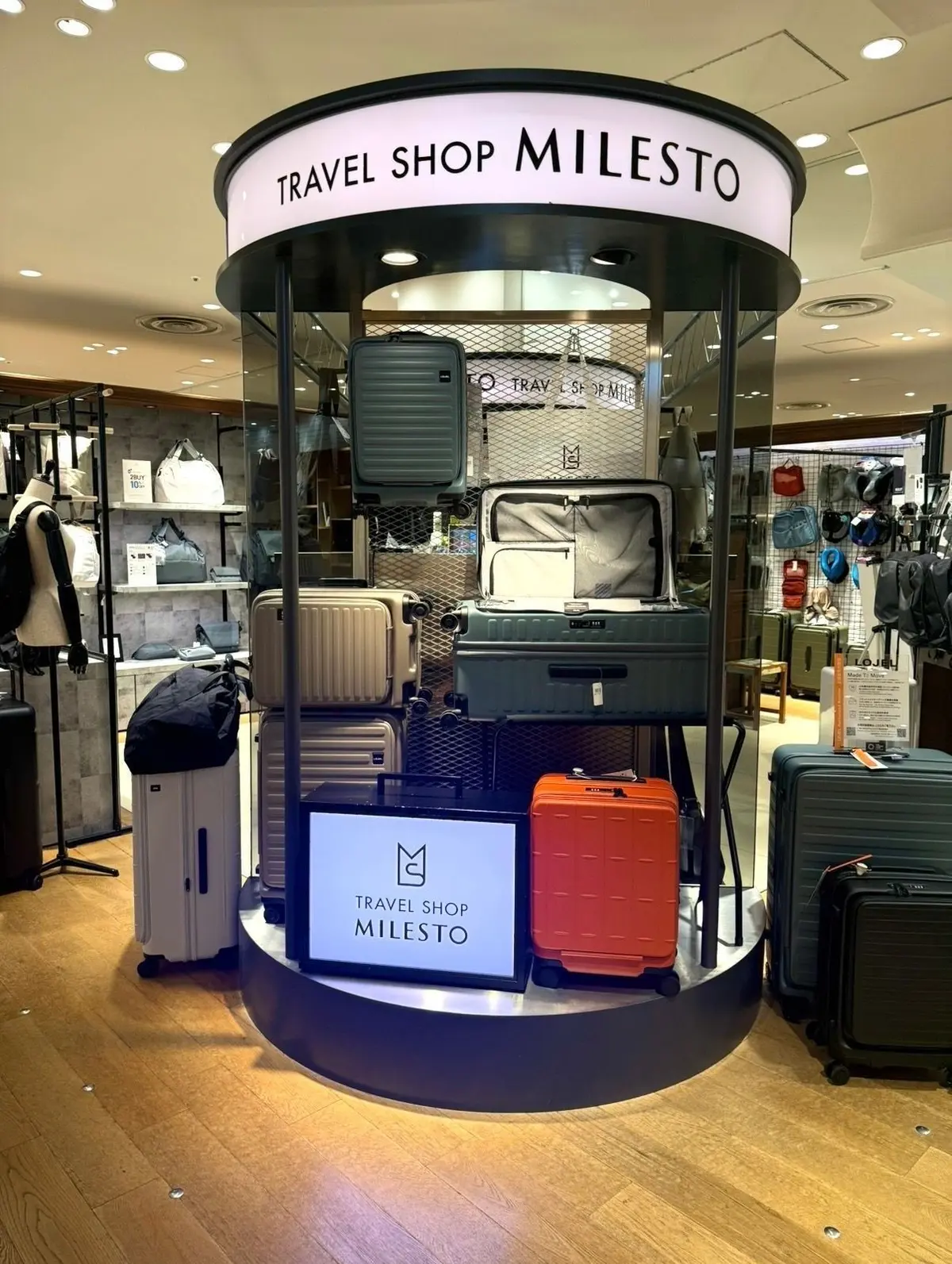 TRAVEL SHOP MILESTOの円形ディスプレイに、グレー、ベージュ、オレンジ、黒の旅行用スーツケースやリュックが展示されている。