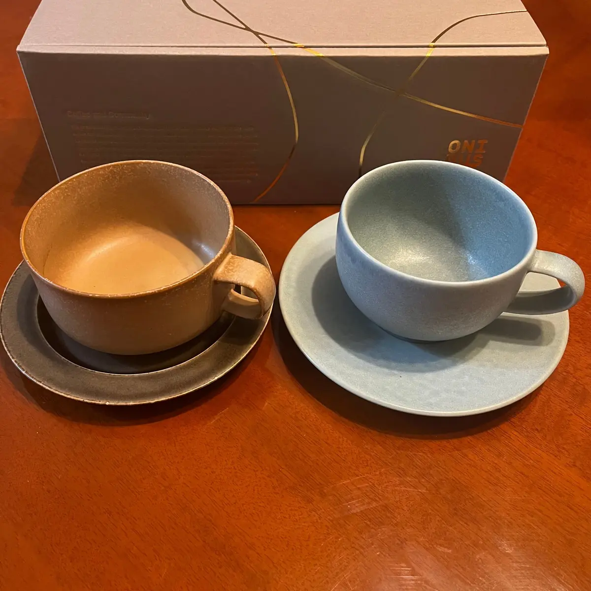 木製のテーブルに置かれた、茶色と水色の陶器のカップとソーサーのセット。カップにはそれぞれコーヒーと水が注がれている。背景には、ONIというロゴが入った白い箱。