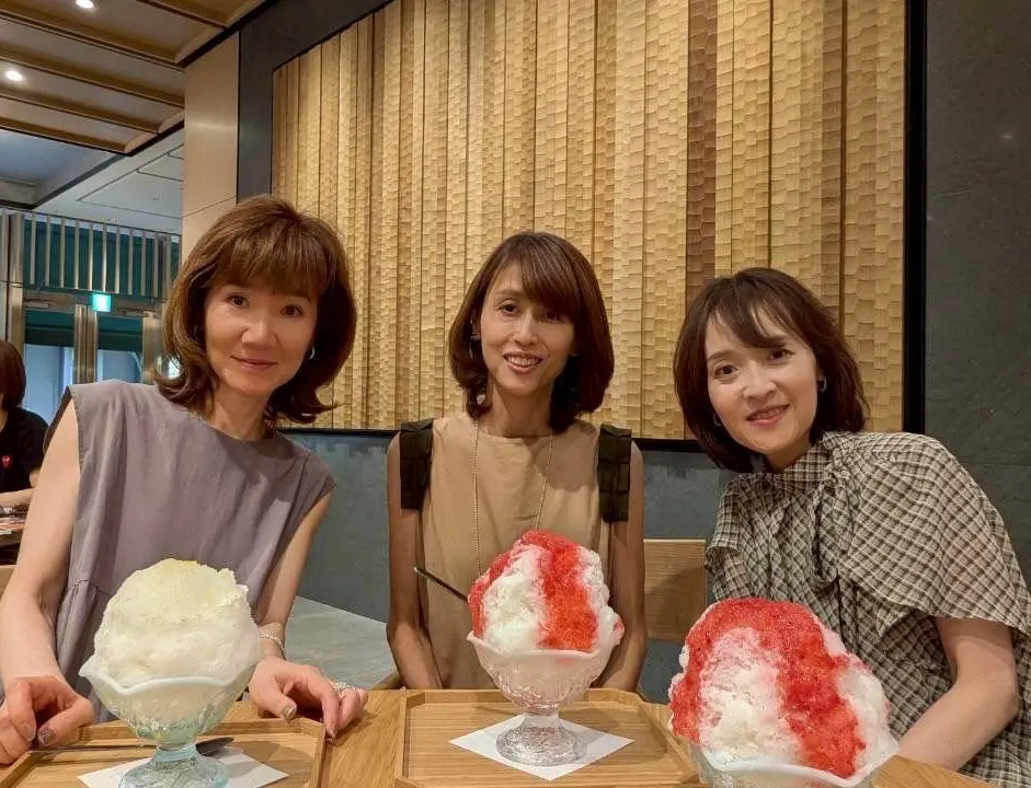 笑顔の女性3人が、かき氷を前にテーブルを囲んでいる屋内写真。左から茶色の髪の女性、ベージュのトップスを着た女性、チェック柄のシャツを着た女性。背景には木目調の壁と照明が見える。