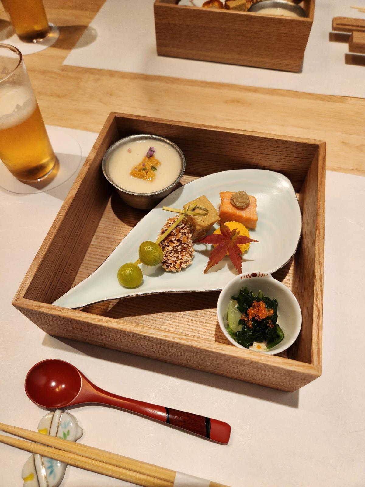 木製の長方形のトレイに盛り付けられた、日本料理のコース料理。葉の形をした白い皿に、様々な食材が彩り豊かに並び、小さな器には海藻と魚卵の和え物が。背景には飲み物と、木製の小箱に入ったお菓子が見える。