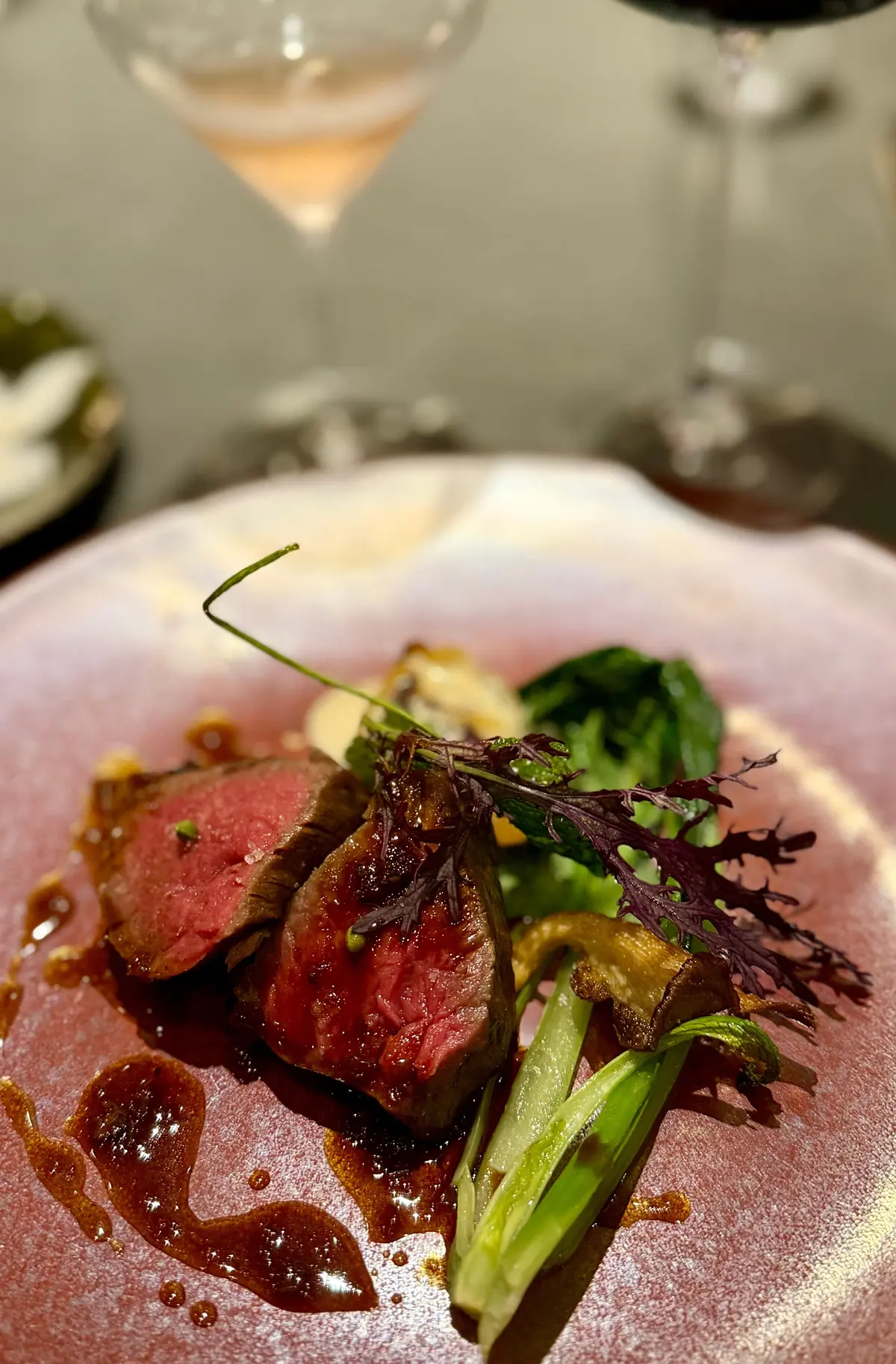 ピンク色の皿に盛り付けられた牛肉料理。赤身の肉は断面がピンク色で、ソースがかけられている。緑色の野菜と、赤い葉の飾り付けが添えられている。背景にはワイングラスが見える。
