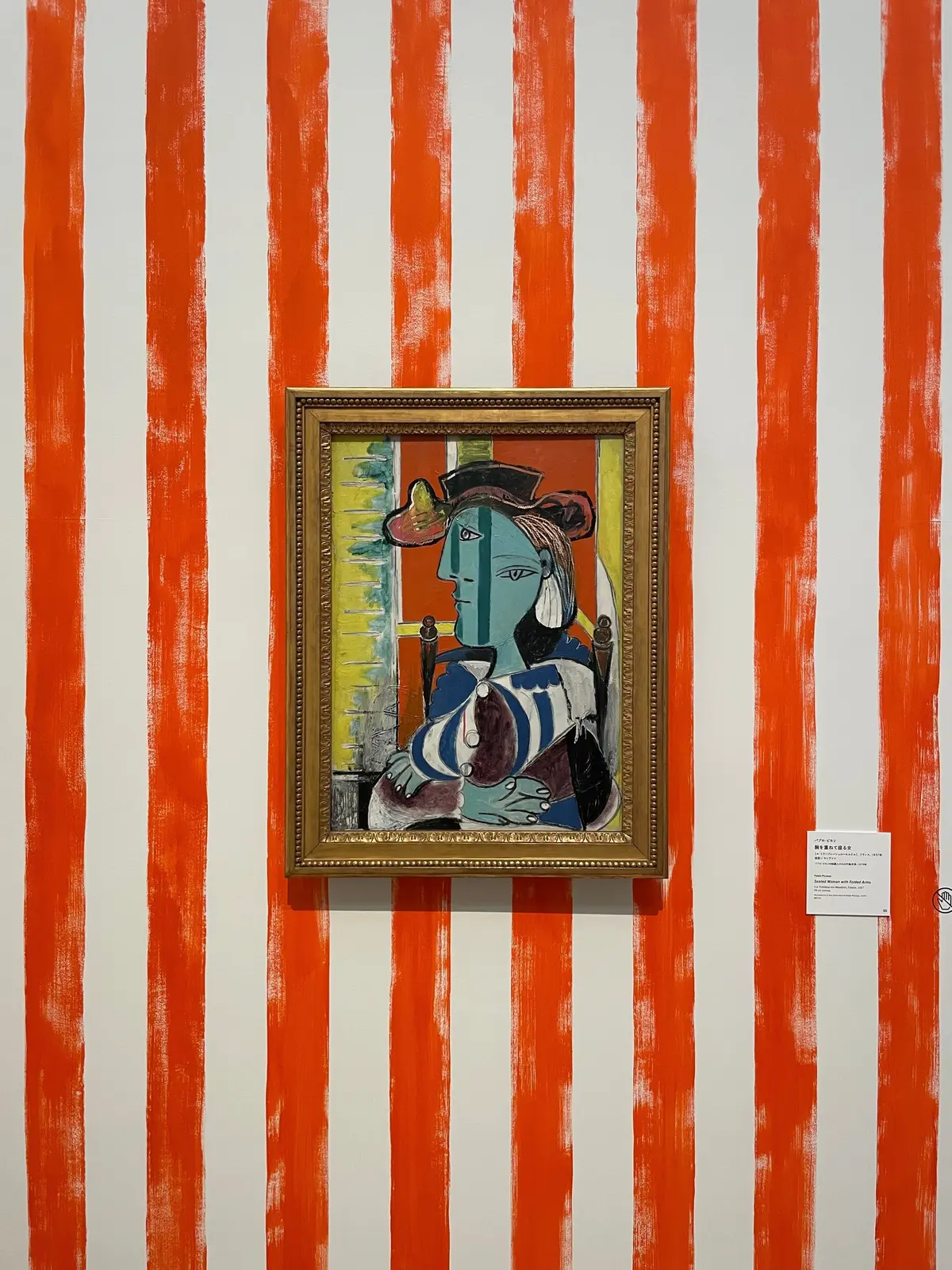 青い肌の女性が帽子をかぶり、青と白の服を着た絵画が金色の額に入れられ、赤と白のストライプ模様の壁に飾られている