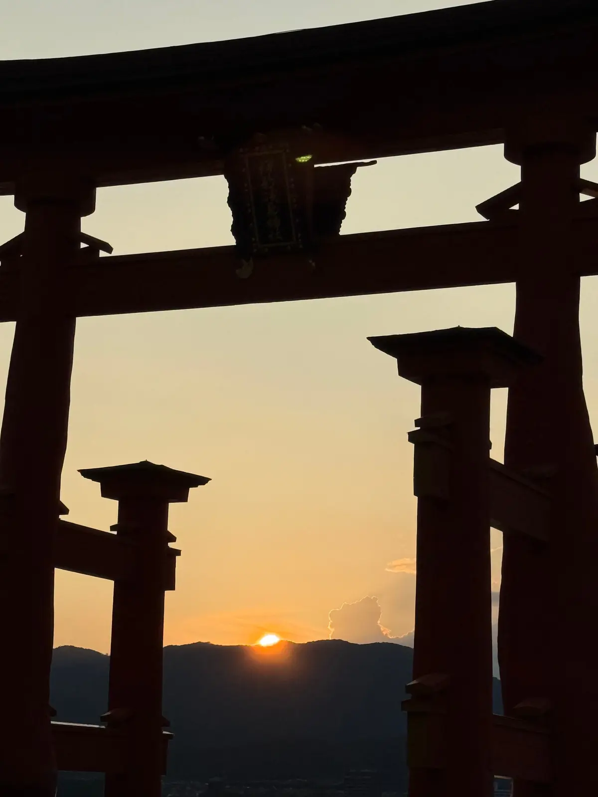 夕焼けを背景にした、朱色の鳥居。中央に太陽が沈み、遠くの山並みと雲が見える。鳥居には額がかけられている。