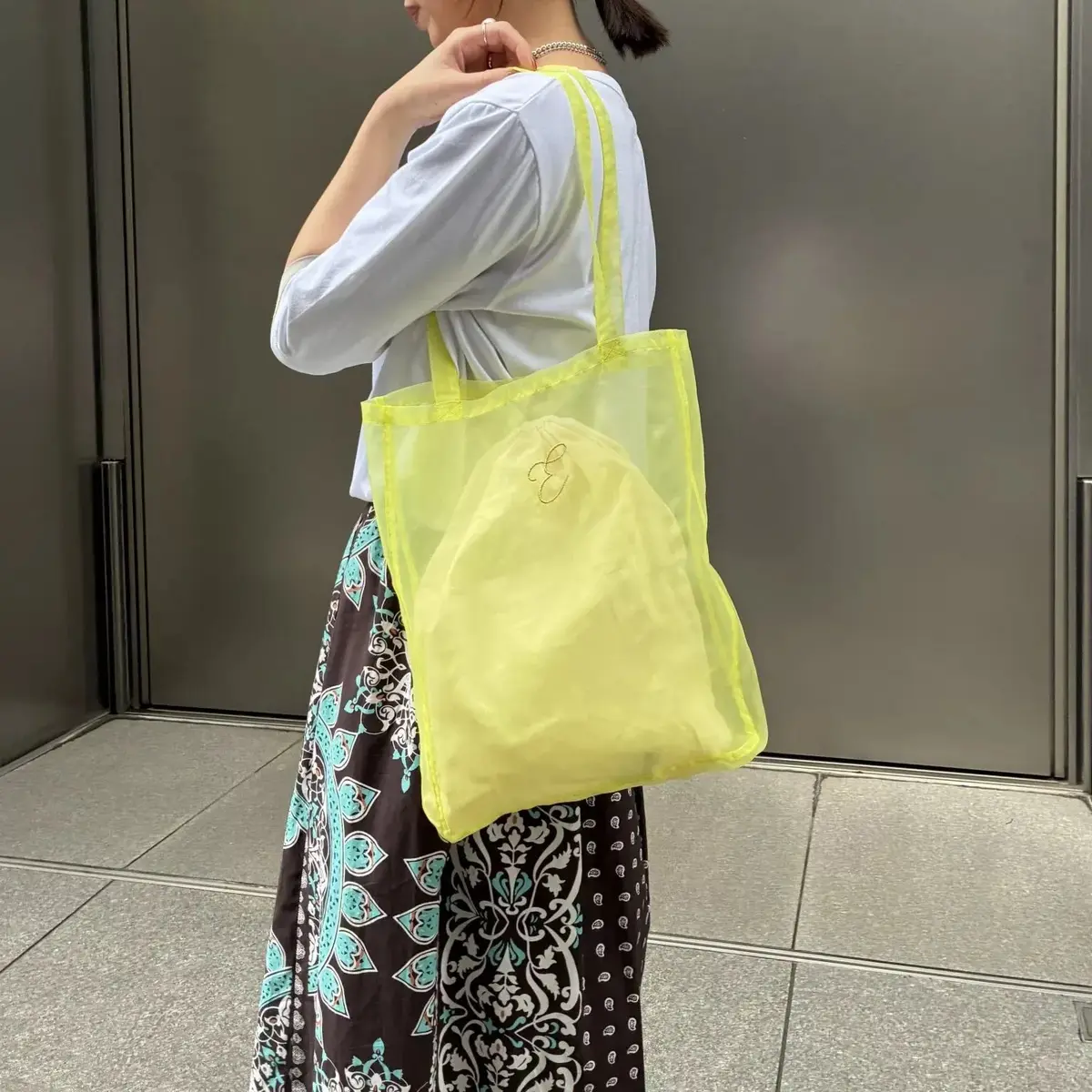 明るい黄色メッシュトートバッグを背負った女性で、建物のガラスドアの前で撮影。女性は白い半袖シャツと花柄スカートを着用。