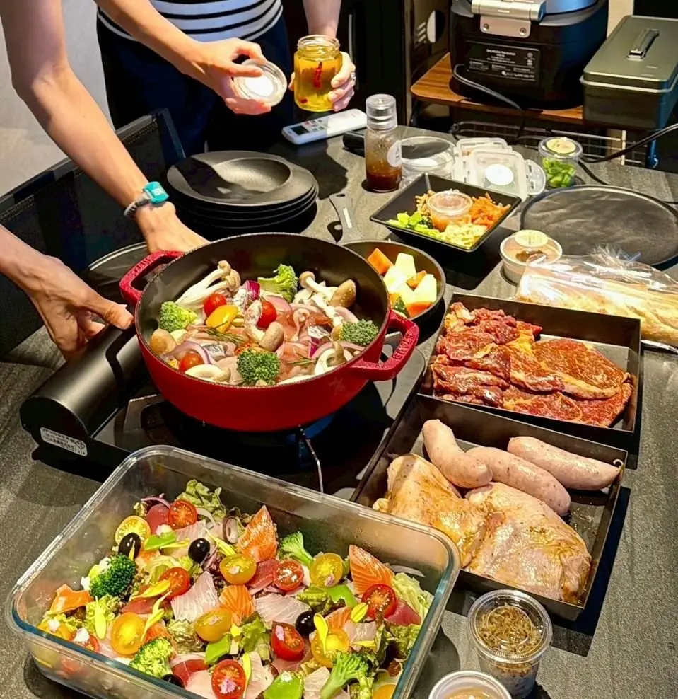 赤色のスキレットで野菜炒め、周囲に肉やサラダ、調味料が並んだ食卓。女性が調味料を注いでいる。