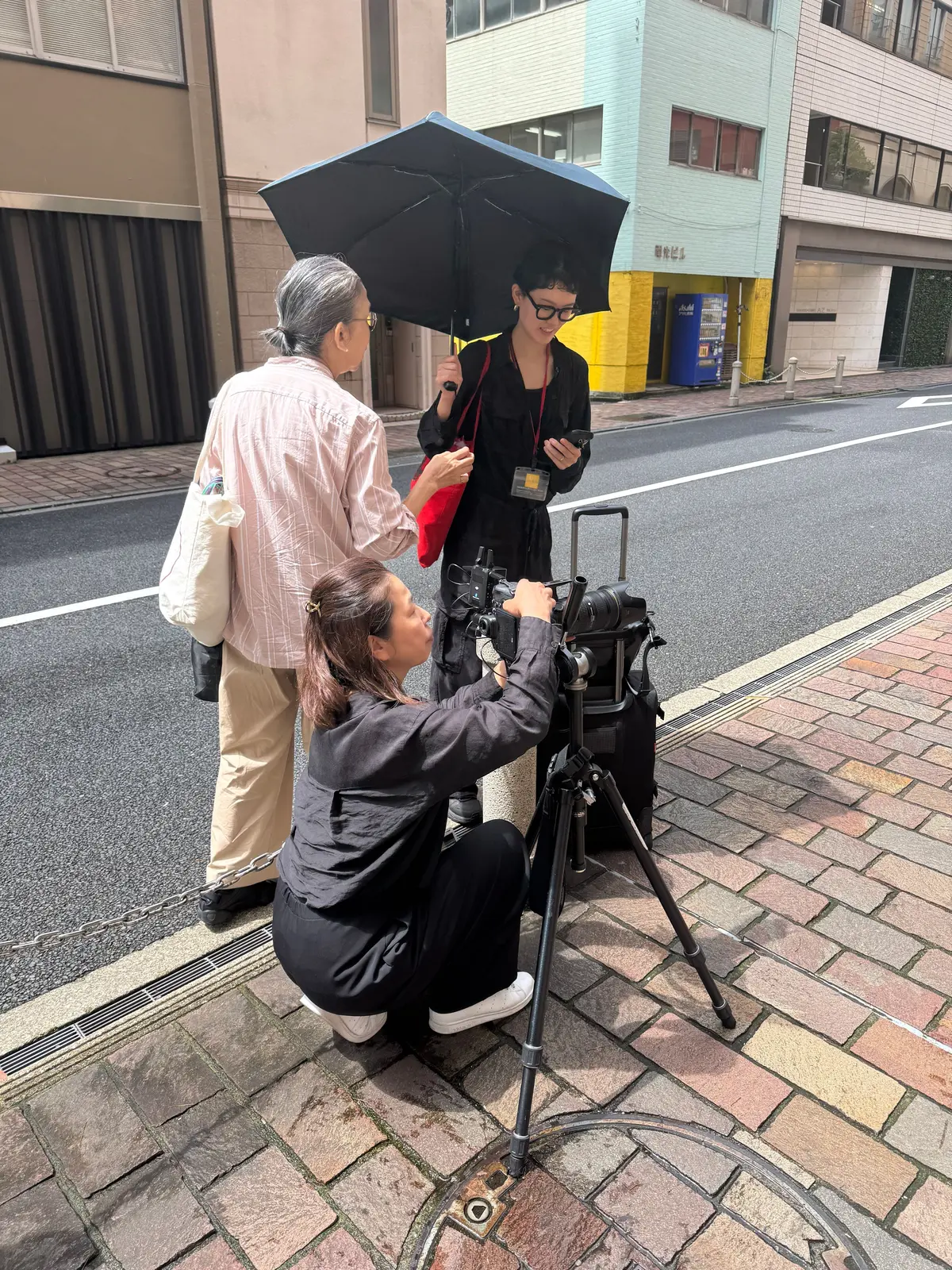 路上で写真撮影が行われている様子。女性カメラマンが三脚を設置し、大型カメラで撮影している。黒い服を着た女性が傘をさし、高齢の男性が立っている。背景には、淡い緑色の建物と自動販売機が見える。