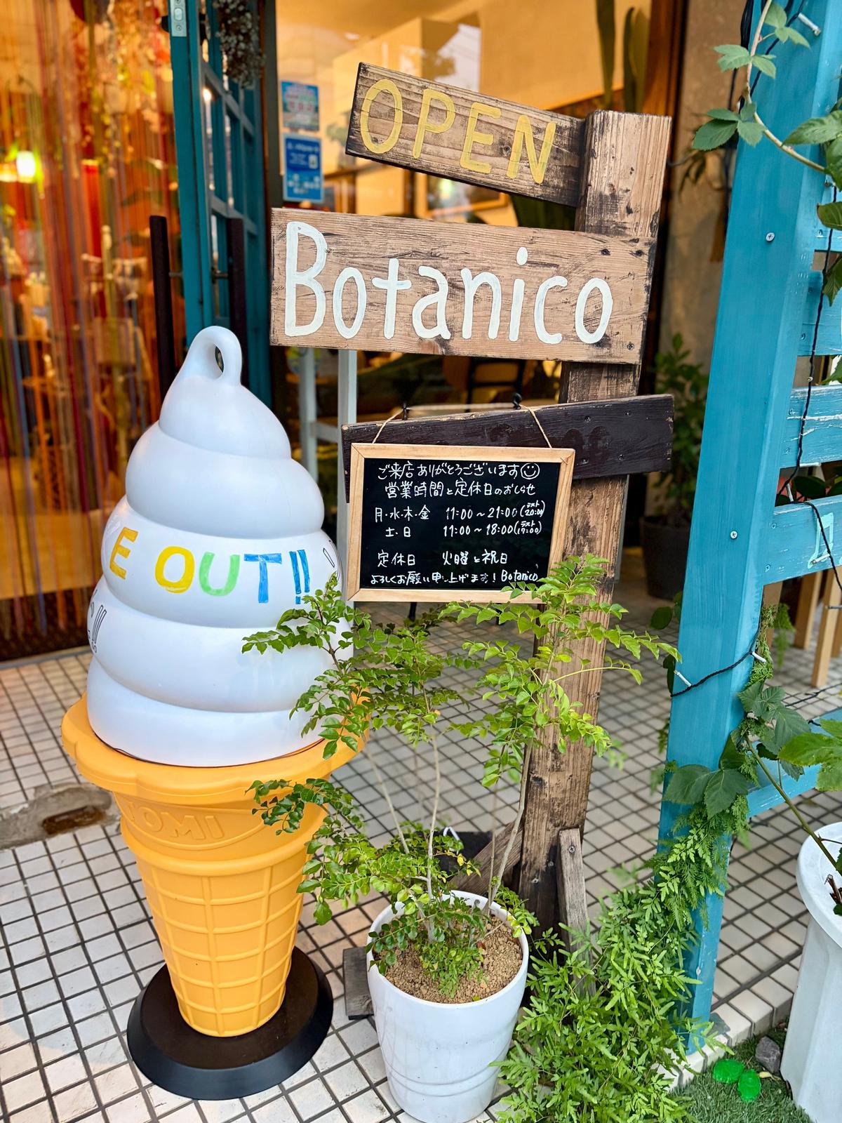 青と白のドアに「Botanico」と書かれた看板があり、その前にアイスクリームの置物と植木鉢に入った植物が置かれている。背景には石畳の道と山が見える。