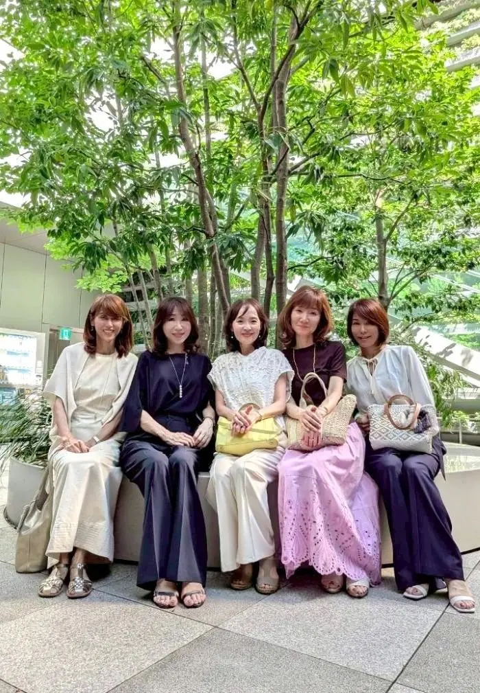 5人の女性が並んで座り、笑顔でカメラに向かっている。手にはさまざまな色のハンドバッグを持ち、自然な緑の背景が広がっている。