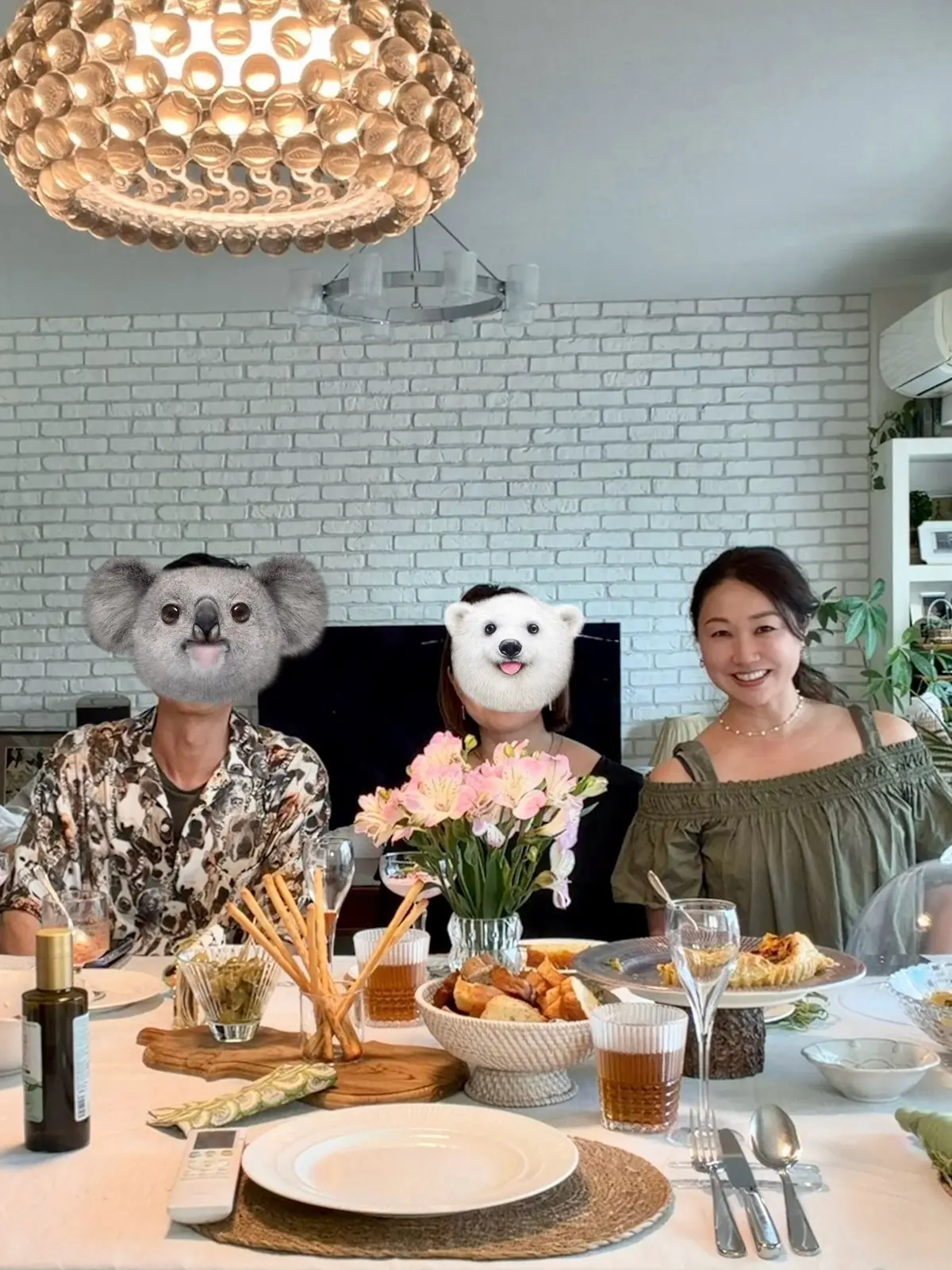 テーブルに料理と花が置かれたリビングで、3人の人物が座っている。中央にピンクの花の花瓶があり、周囲にはパスタ、パン、ワインが並ぶ。背景には白いレンガの壁とモダンな照明が設置されている。