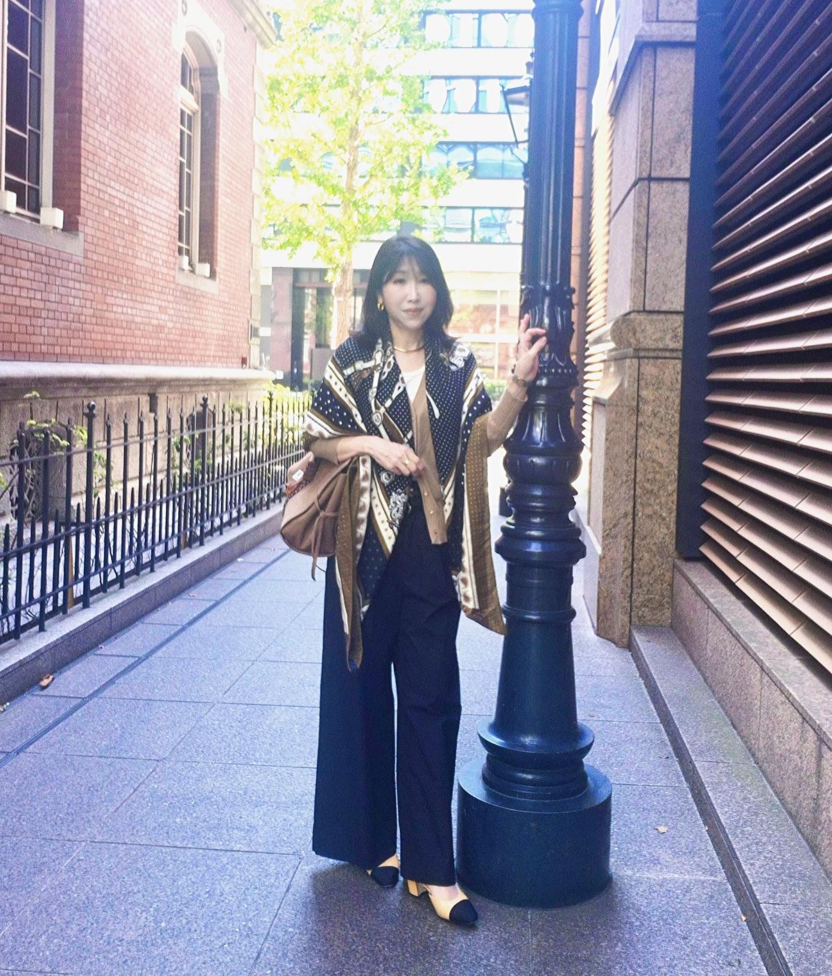歩行中の女性。黒いパンツと茶色のトップスに、柄のショールとスカーフを着用。茶色のバッグを肩にかけ、黒いパンプスを履いている。背景はコンクリートの建物と通路。
