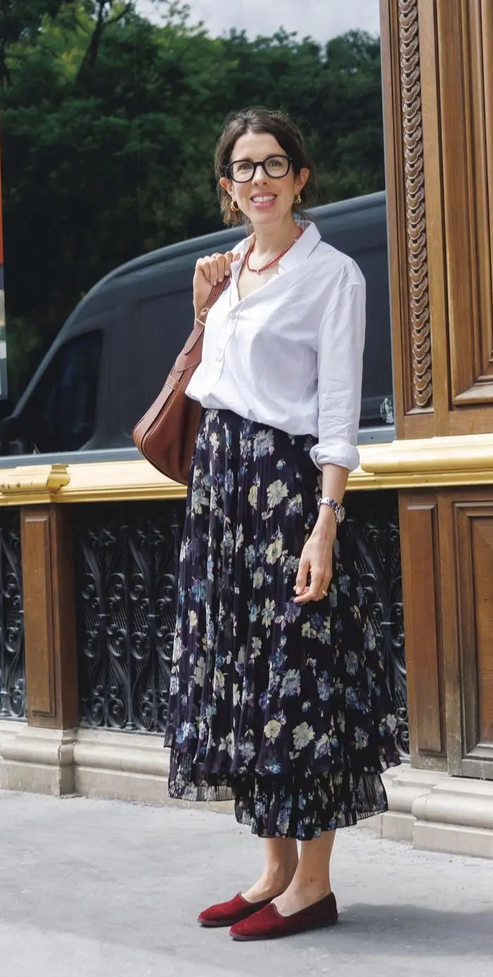 白シャツに花柄のフレアスカート、赤いパンプスを履いた女性。眼鏡をかけ、茶色の肩掛けバッグを持つ。背景には木や建物がある。