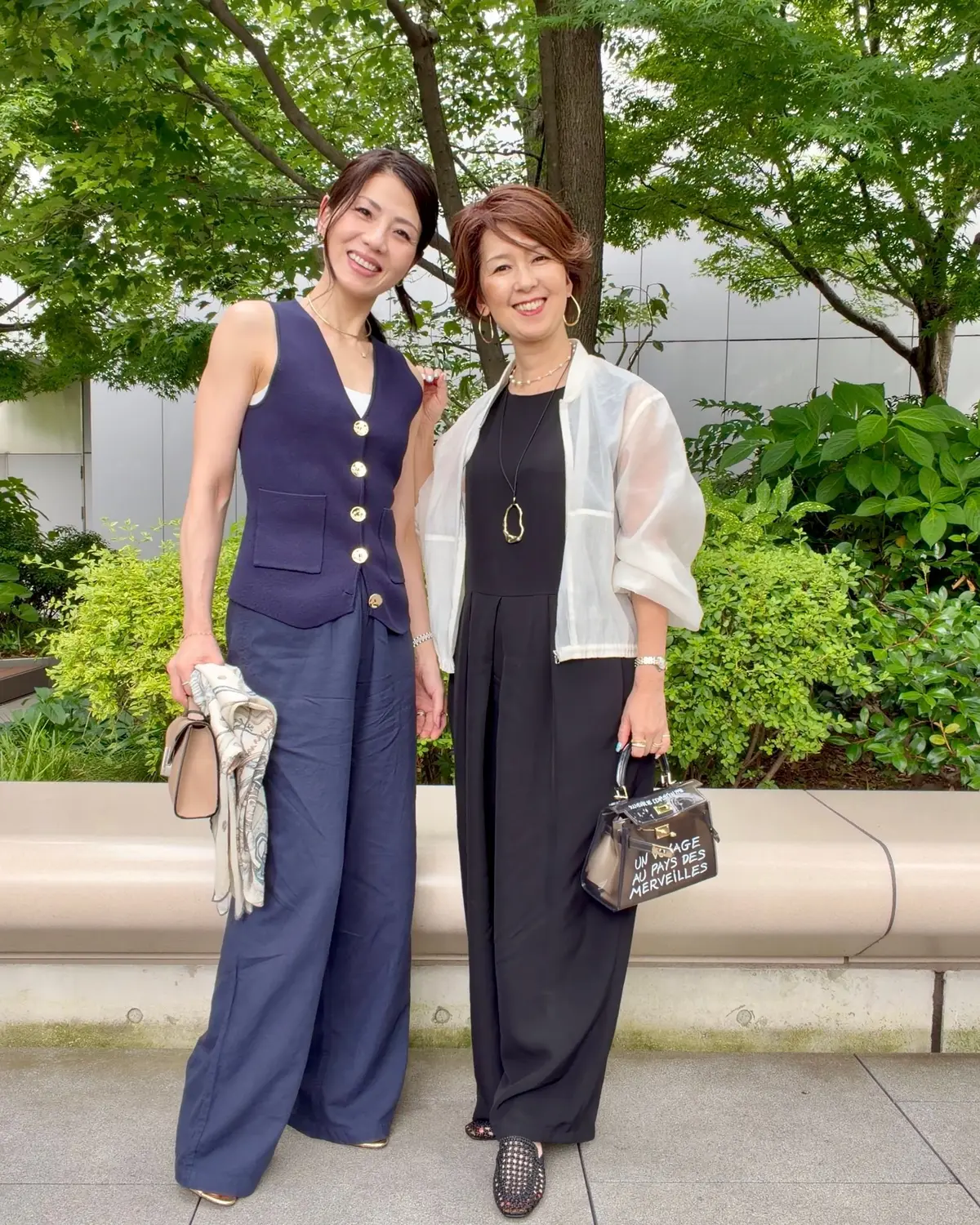 二人の女性が屋外で並んで立っている。左の女性は紺色のベストとワイドパンツを着て、右の女性は黒のワンピースに白のシースルーカーディガンを着用。両方とも笑顔で、背景には緑の木や低木が広がっている。