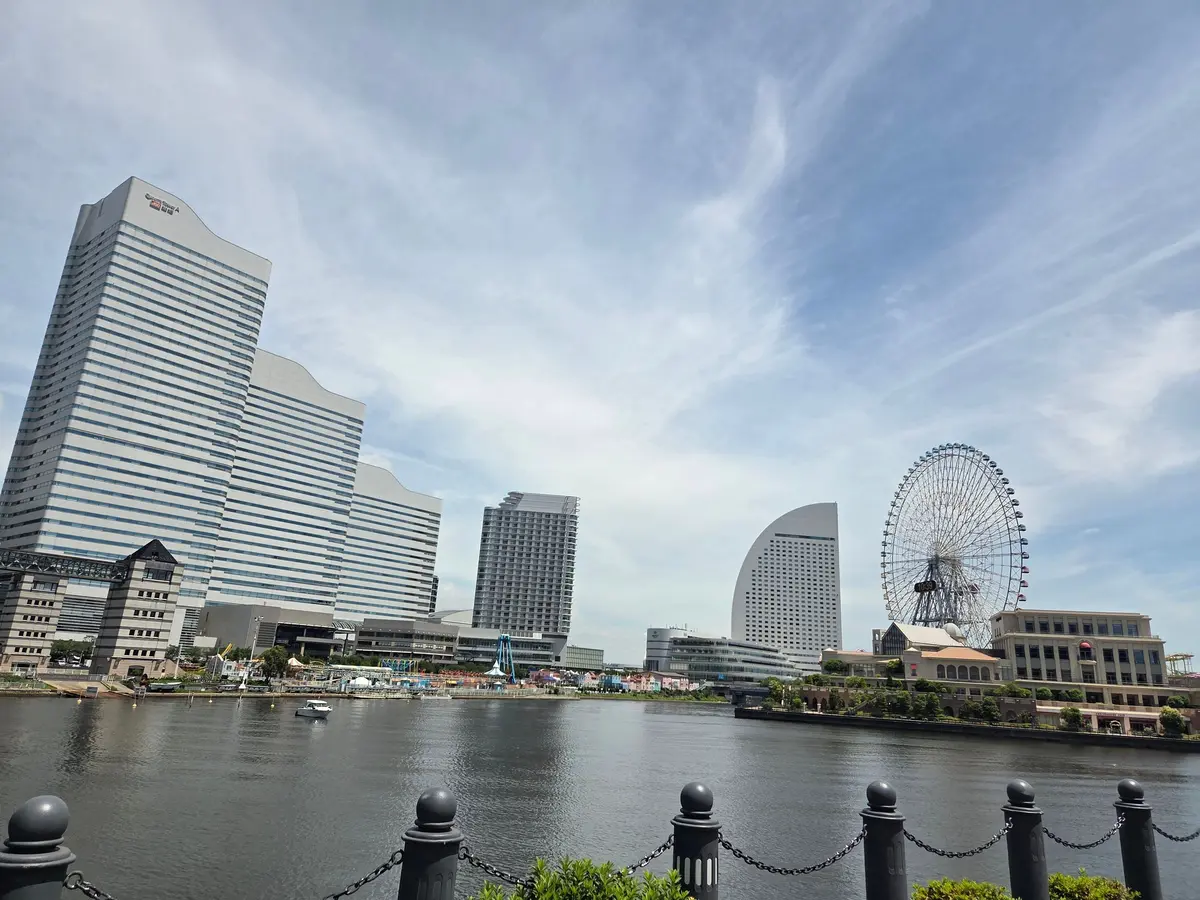 横浜の港町風景。水辺に高層ビル群と観覧車が並び、晴天の青空が広がる。左には大型のオフィスビル、右には観覧車が目立つ。