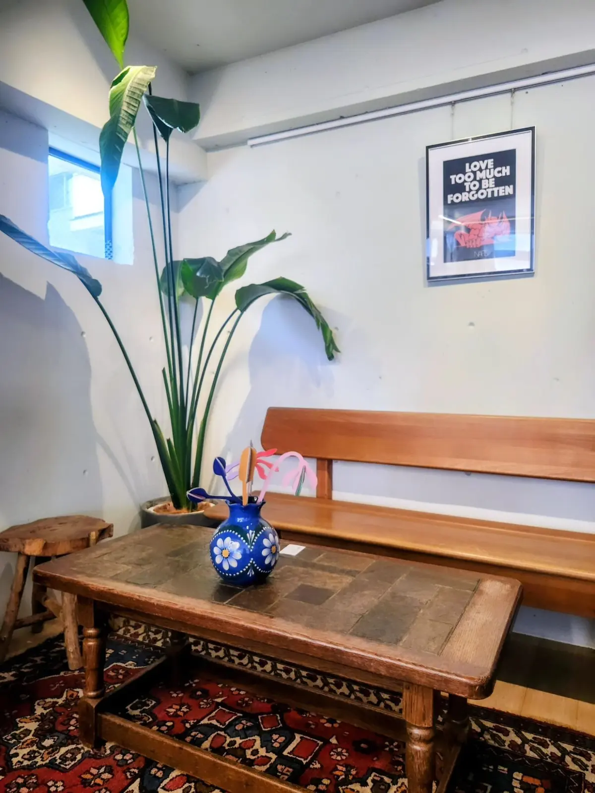 木製のコーヒーテーブルに、青い花瓶に入った白い花とピンクのオブジェが置かれている。背景には窓とベンチ、そして「Love too much to be forgotten」と書かれた絵画がある。テーブルの下には赤い絨毯が敷かれている。