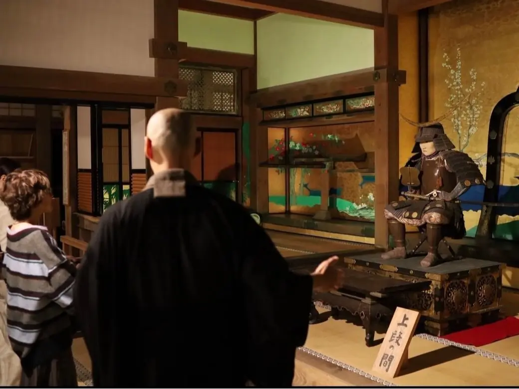 伝統的な日本の部屋で、武者人形が座っている。正面には黒装束の男性が説明をしている様子。背景には障子や襖、花瓶などが配置されている。