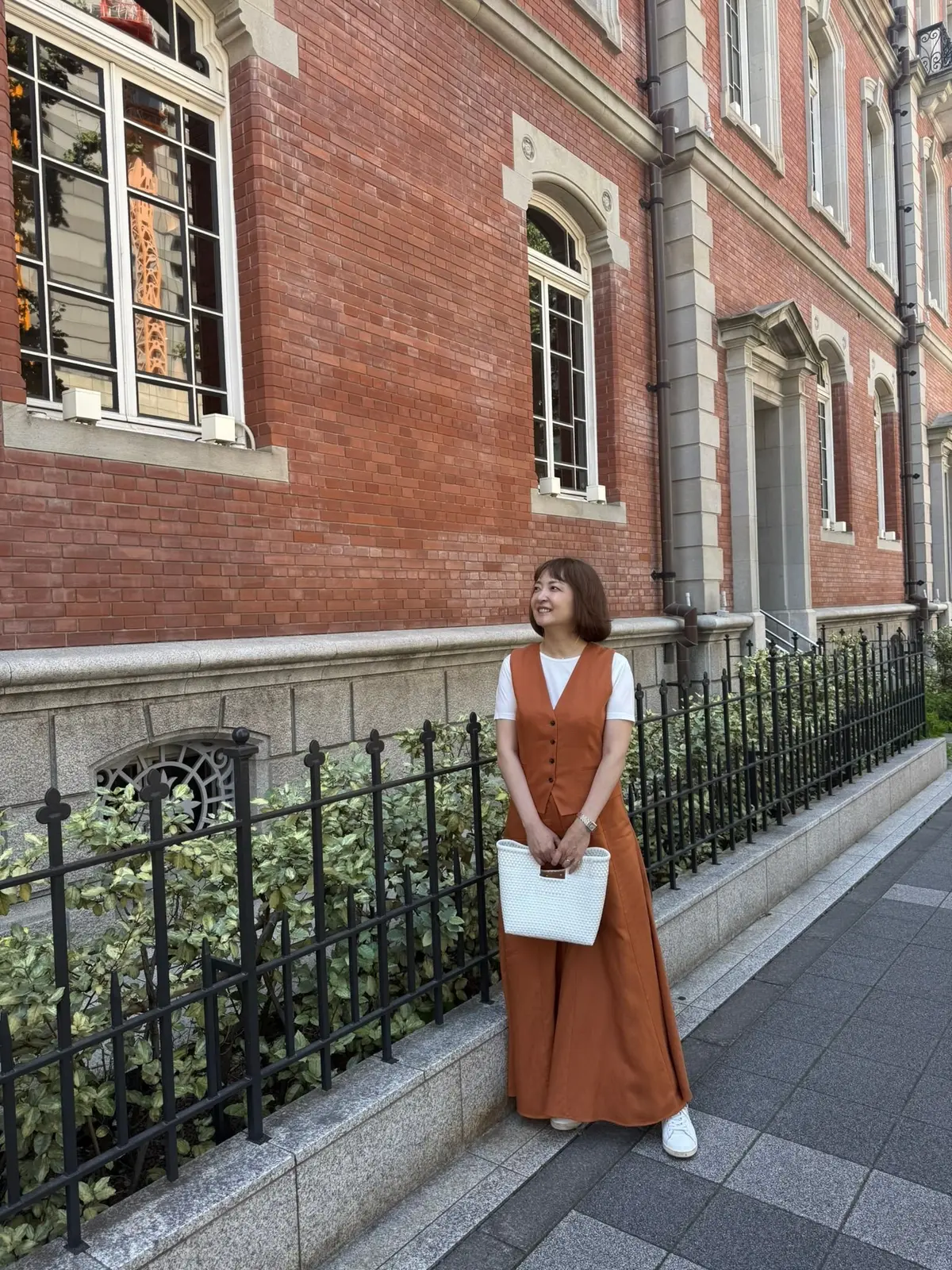 茶色のオーバーオールとロングスカートを着用した女性が、鉄柵のそばに立ってこちらを見ている。背景にはレンガ造りの建物と白い窓枠が見える。