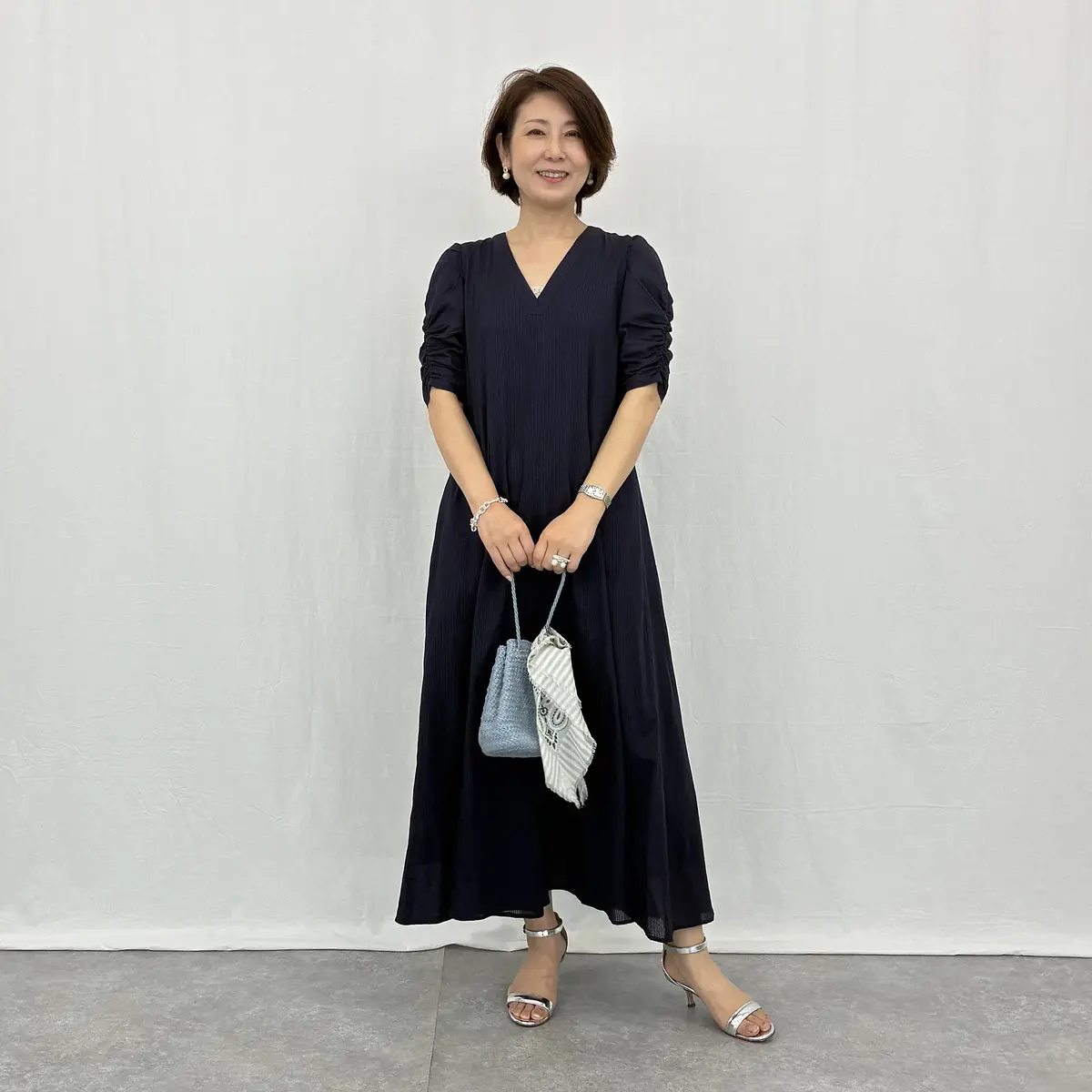 紺色のAラインワンピースを着た女性。Vネックで、長袖で袖にボリュームがある。シルバーのサンダルを履き、手には青いバッグを持っている。笑顔で正面を見ている。