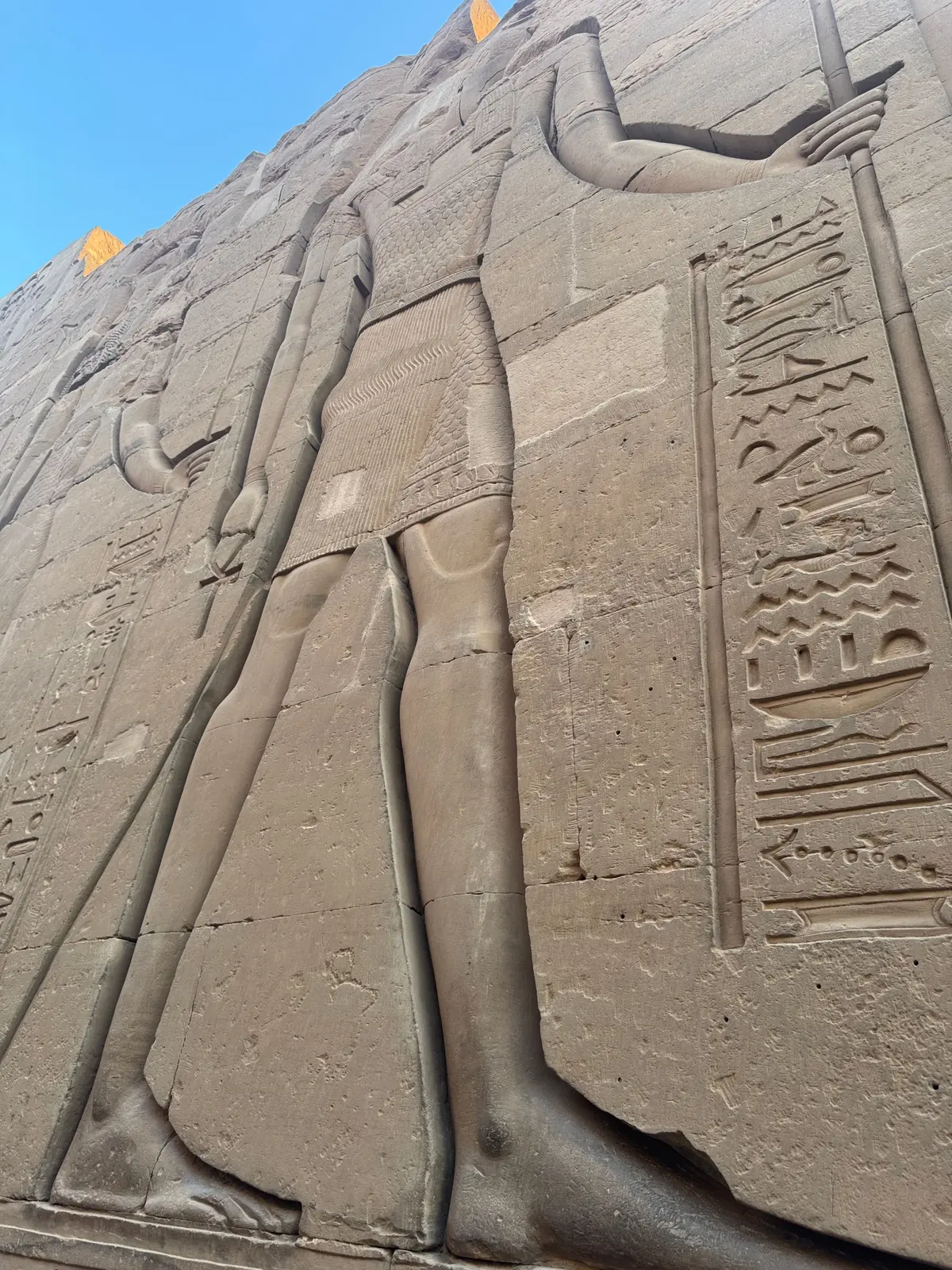 古代エジプトの神殿壁に彫られた巨大な石像群。男性像が直立し、腕を上げている。壁面にはヒエログリフが刻まれている。背景は青空。