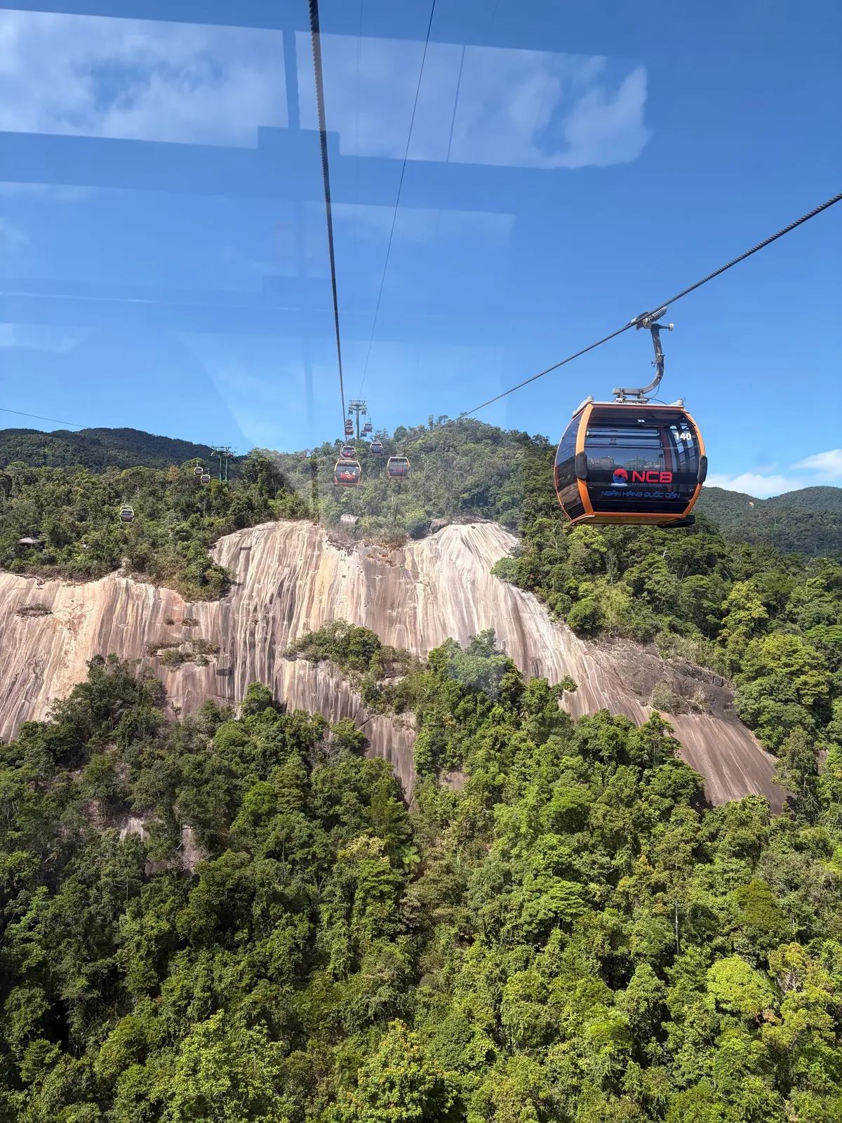 緑豊かな山々と白い岩壁の上を、Nami Island Cable Car（南怡島ケーブルカー）のゴンドラが運行している様子。青空に白い雲が浮かび、背景には緑の山々が連なっている。