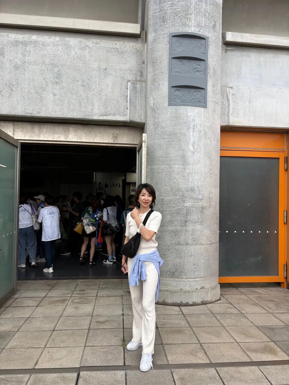コンクリート柱の横に女性が立ち、ベージュの服と白いスニーカーを着用。手には黒いショルダーバッグをかけ、青いスカーフを首に巻いている。背景には建物の入り口から人が入り、群れで待っている様子が見える。