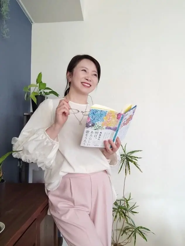 女性が白いブラウスとピンクのズボンで本を読み、メガネをかけた笑顔の姿。背景には観葉植物と青い壁がある。