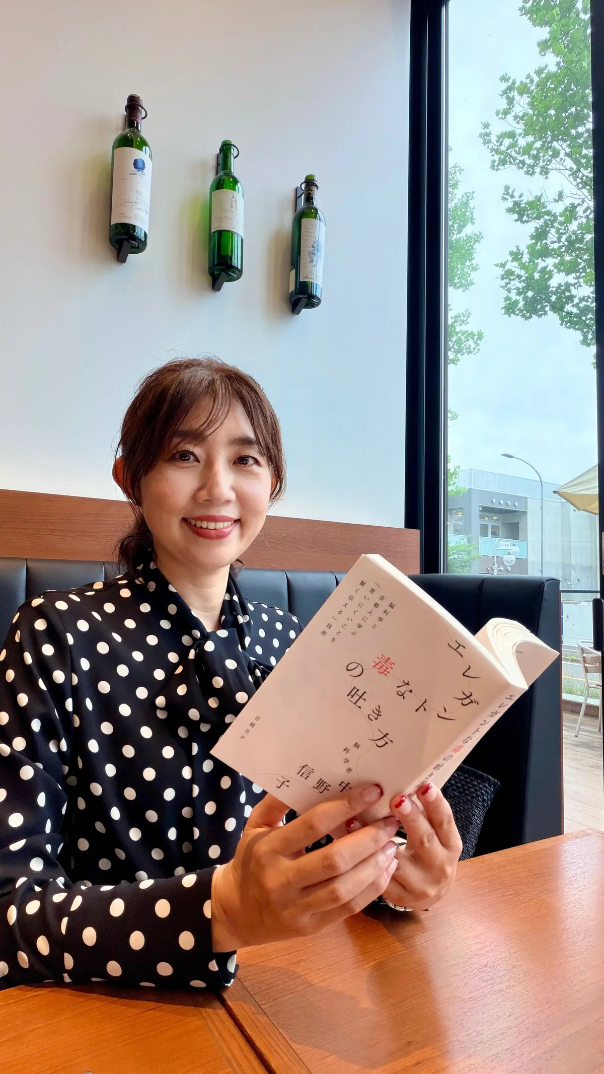 女性が黒のドット柄の服を着て、木製のテーブルに座り、日本語の本を手に持つ。背景には窓から外の木々が映り、壁には緑のボトルが飾られている。