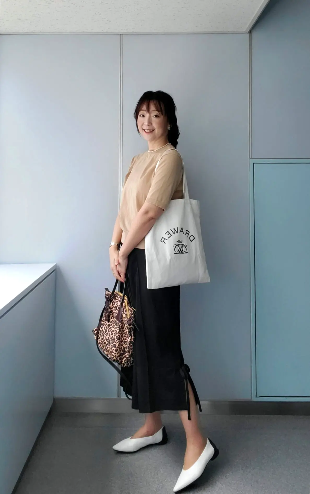 ベージュのトップスと黒いパンツルーズフィットの女性。白のキャンバストートバッグを持ち、茶色のレオパード柄ショルダーバッグを肩に掛けている。白いフラットシューズを履き、背景は淡いグレーと白の壁。