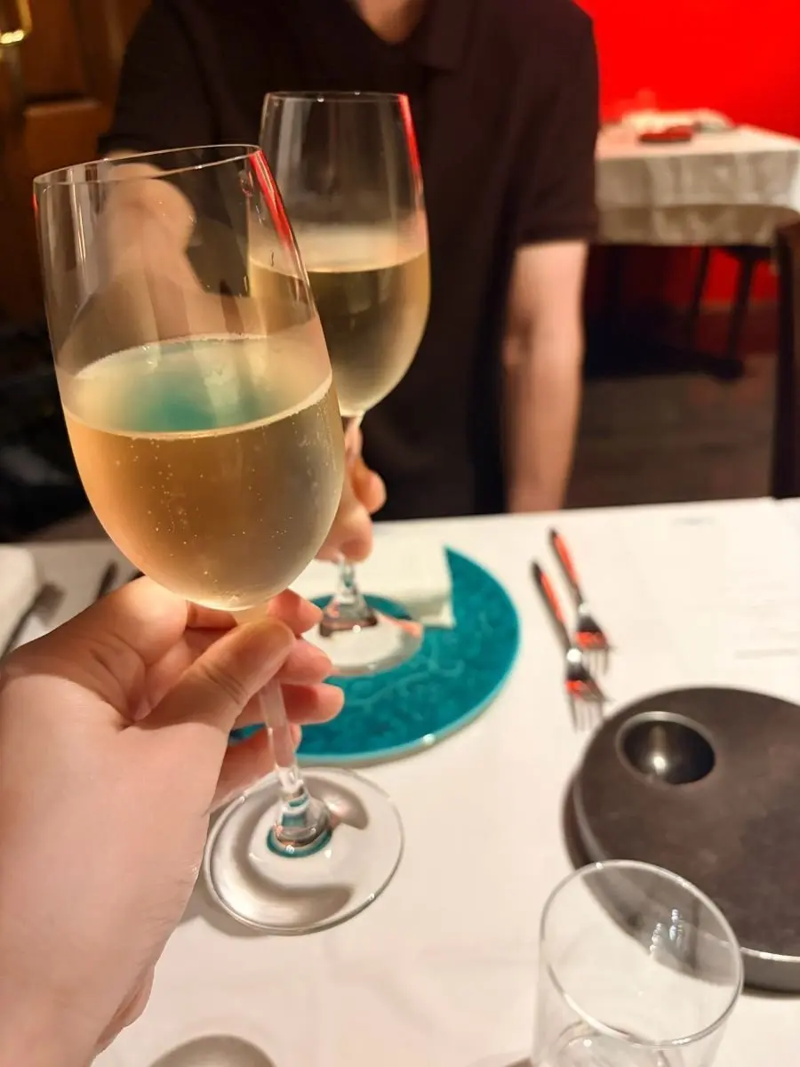 白ワインのグラス2つが触れ合って、乾杯している様子。テーブルには青色のコースターとカトラリーが置かれている。背景には人物と赤い照明が見える。