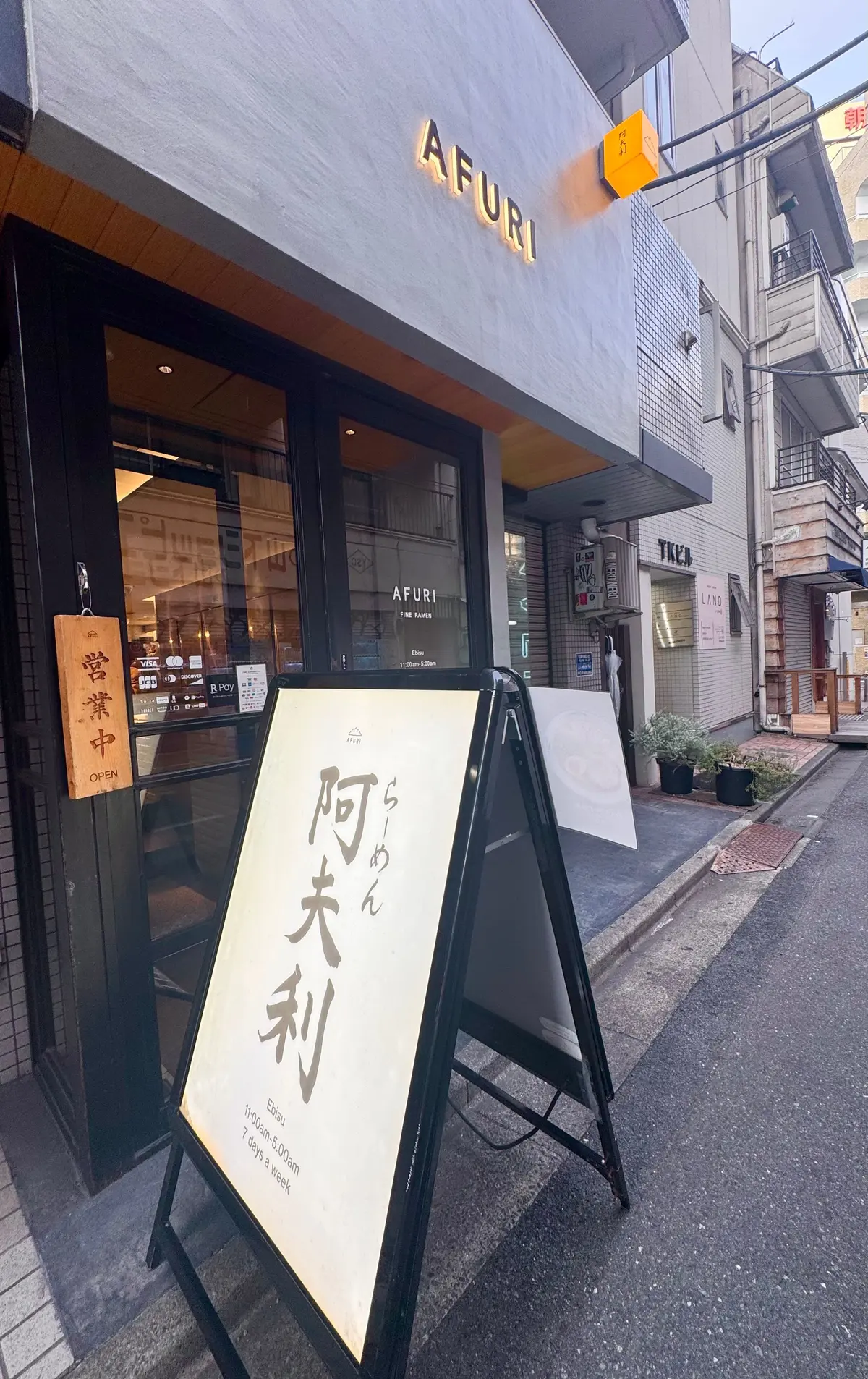 AFURIのラーメン店外観。グレーの建物で、大きなガラス戸を開け、店内が見える。看板には「阿夫利」の文字と営業時間が記載。背景には、路地裏の他の店舗が並ぶ。