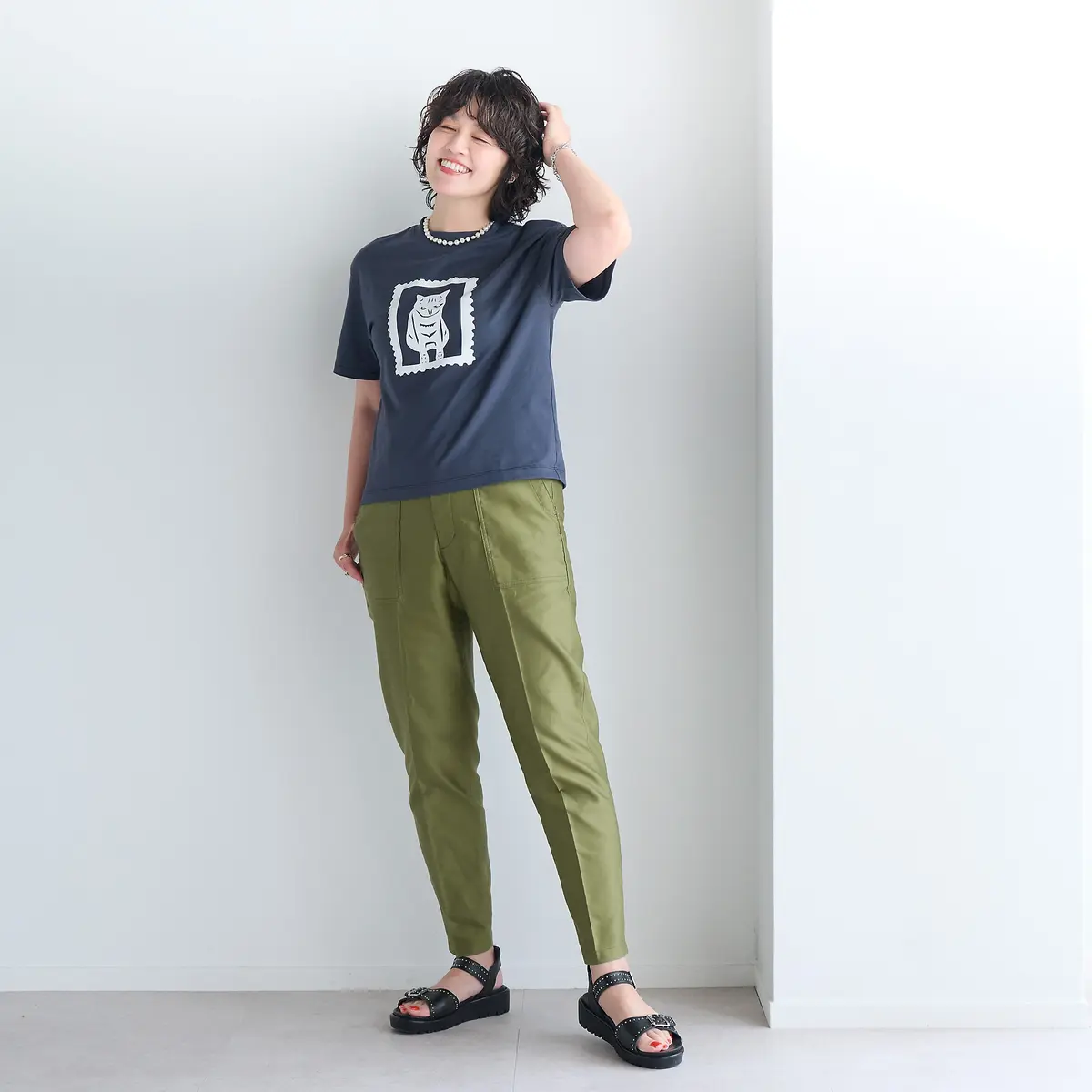 白い壁の前に立つ女性。ネイビーの猫のイラストがプリントされた黒いTシャツと、カーキ色のテーパードパンツを着用。黒いサンダルを履き、片手を頭に当てて笑顔。白い真珠のネックレスを身につけている。