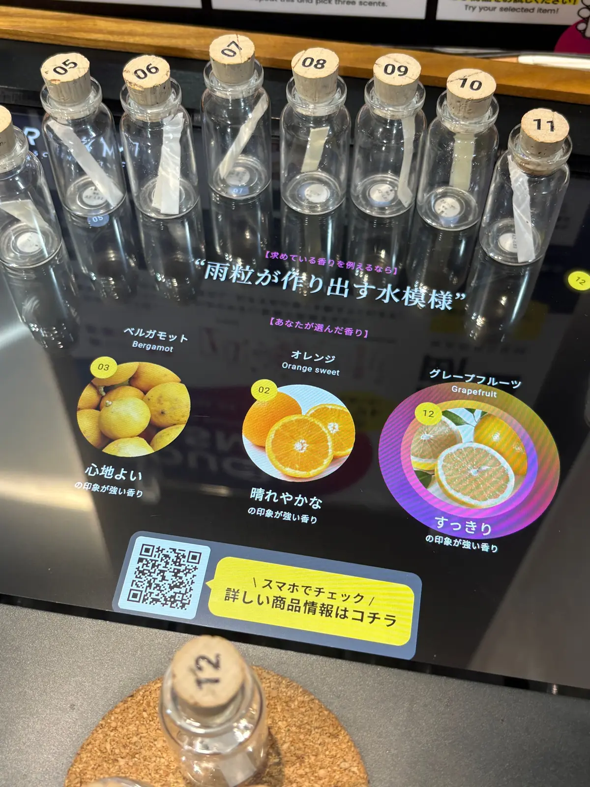 12本のガラス瓶が並び、番号付きコルク栓が装着。レモン、オレンジ、グレープフルーツの画像と香り名が表示された案内板が背景にあり、QRコードと「イスマでチェック」というテキストも見える。