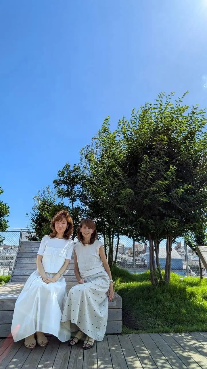 青空の下、白いワンピースを着た2人の女性が、木陰の階段に座っている。左の女性は笑顔で、右の女性は少し控えめな表情。背景には緑の芝生と、遠くの都市の風景が見える。