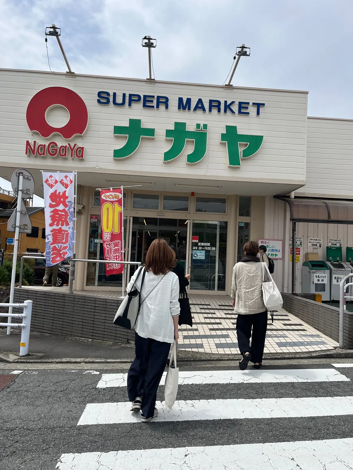 スーパーマーケット「ナガヤ」の入り口で、2人の女性が買い物袋を持って店を出る。建物は白の外壁で、赤と緑のロゴと看板が目立つ。
